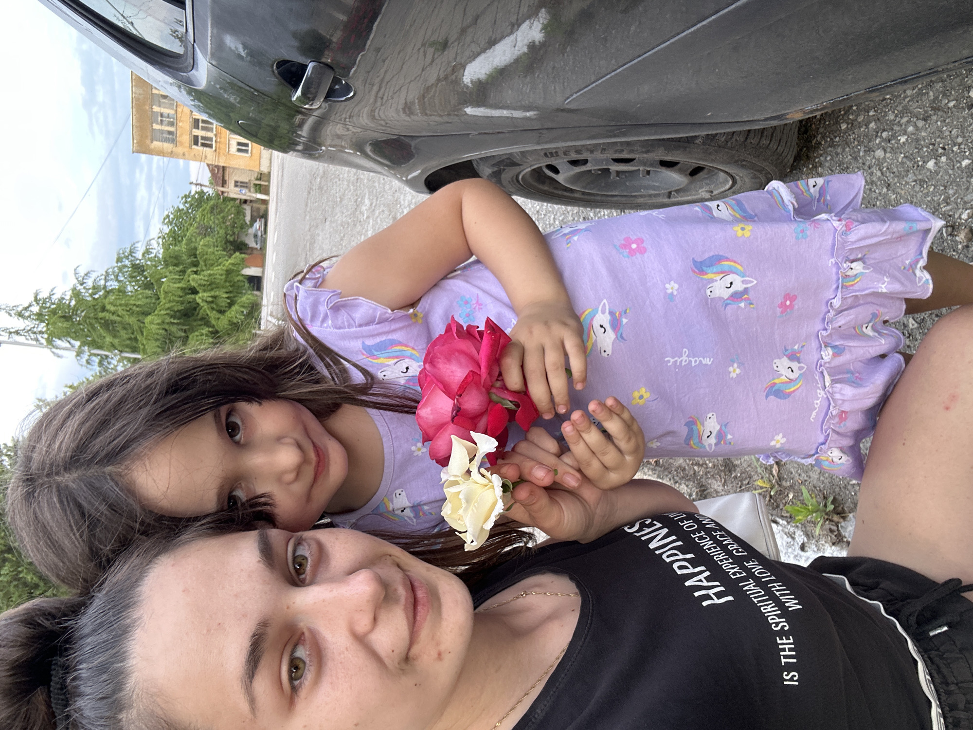 A woman and a young girl are closely posed together in an outdoor selfie taken in Resen, Bulgaria. The image is taken with the lower part of the woman's body visible, and the child positioned above her, partially under the bumper of a parked grey car. The woman is wearing a black t-shirt with the word "Happiness" and a longer phrase in white text, as well as black shorts. The little girl wears a lavender dress with unicorn prints. The two hold a small bouquet of roses, some red and one white. In the background, a light brown building is seen across the road. The ground is concrete.
