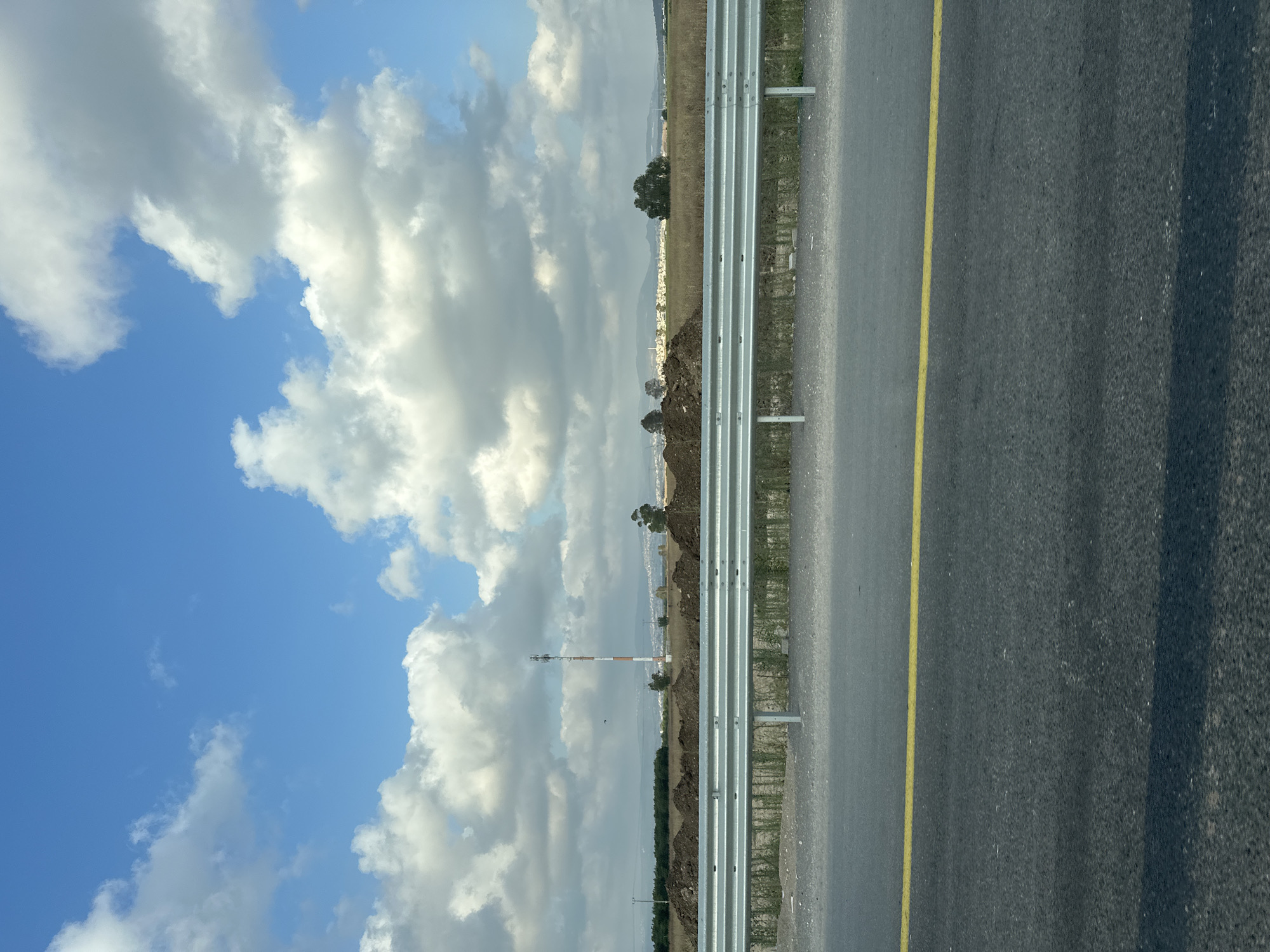 This image depicts a view from a road in Acre, Israel, looking towards a landscape under a cloudy blue sky. In the foreground, a dark asphalt road with a single yellow line runs horizontally across the frame. Beyond the road, a metal guardrail with horizontal bars and vertical supports separates it from a dry, grassy field. Piles of dark soil are visible in the middle ground, suggesting construction or agricultural activity. A tall, slender communication tower, painted with red and white bands, stands prominently in the distance. Further back, a cluster of light-colored buildings forms a town or village. Scattered trees and a line of darker vegetation complete the distant landscape. The overall scene is one of a rural or semi-rural area near the coast, characterized by open spaces, infrastructure, and distant signs of human settlement. There are no people or animals visible in the frame.