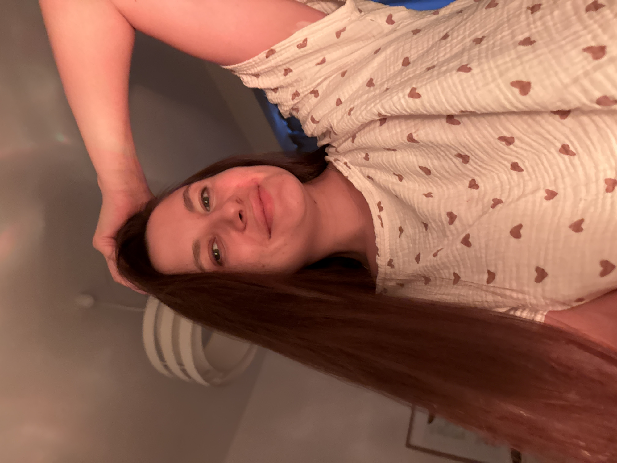 A woman with long, straight brown hair is smiling at the camera. She is wearing a white shirt with small brown hearts. Her left arm is raised above her head. The background is blurry but appears to be an indoor setting with a lamp and a framed picture. The city of Belgrade, Serbia, cannot be determined from the image.