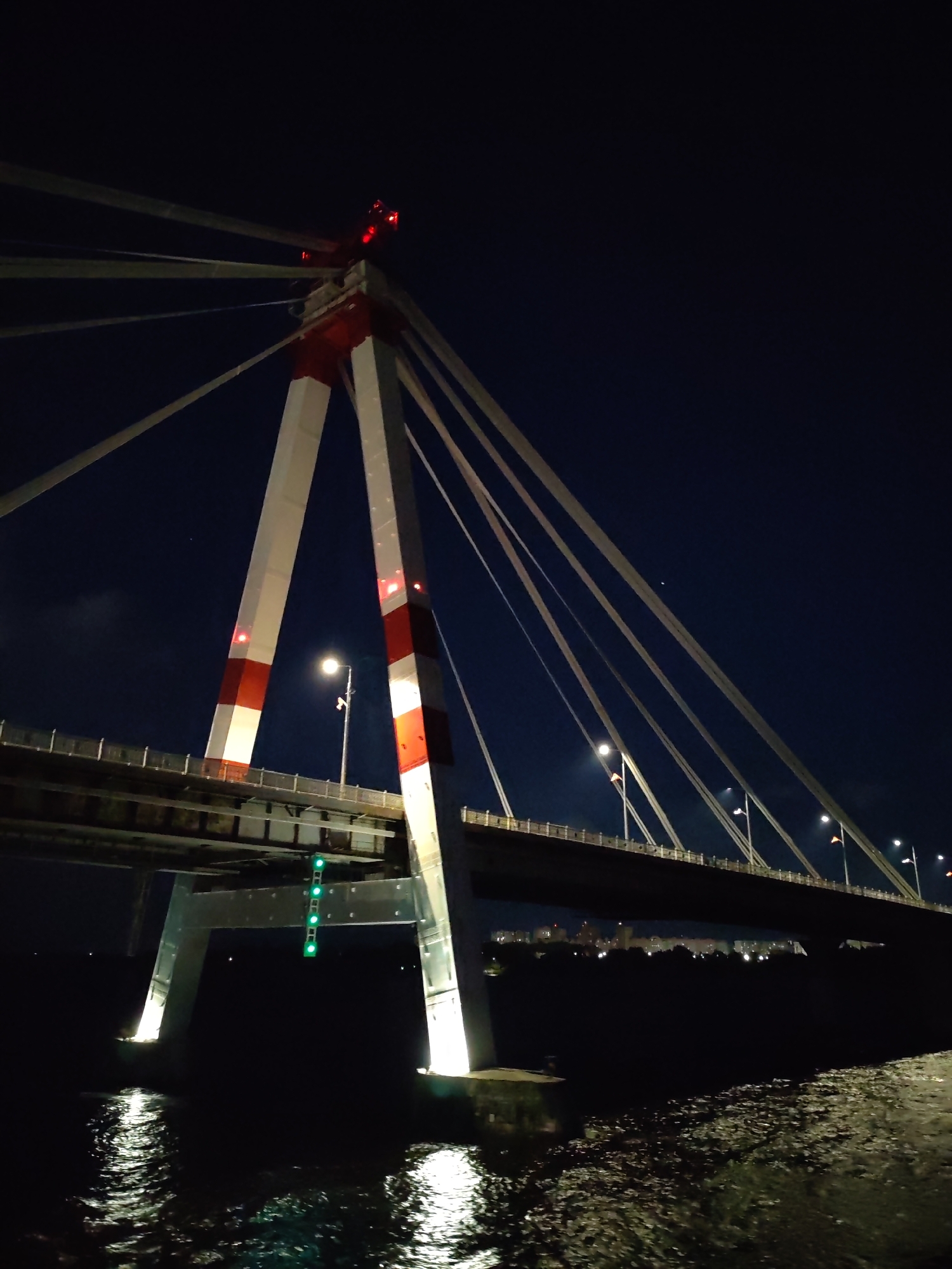 Captured at night, the photograph showcases a bridge in Unknown, Unknown. The bridge, characterized by a central support tower painted white with red sections, is illuminated. Cables extend from the top of the tower. The tower is further marked by red lights at the top. The bridge surface itself is illuminated by streetlights, which are visible along the span. Below the bridge, the water reflects the lights. The sky is a dark blue.