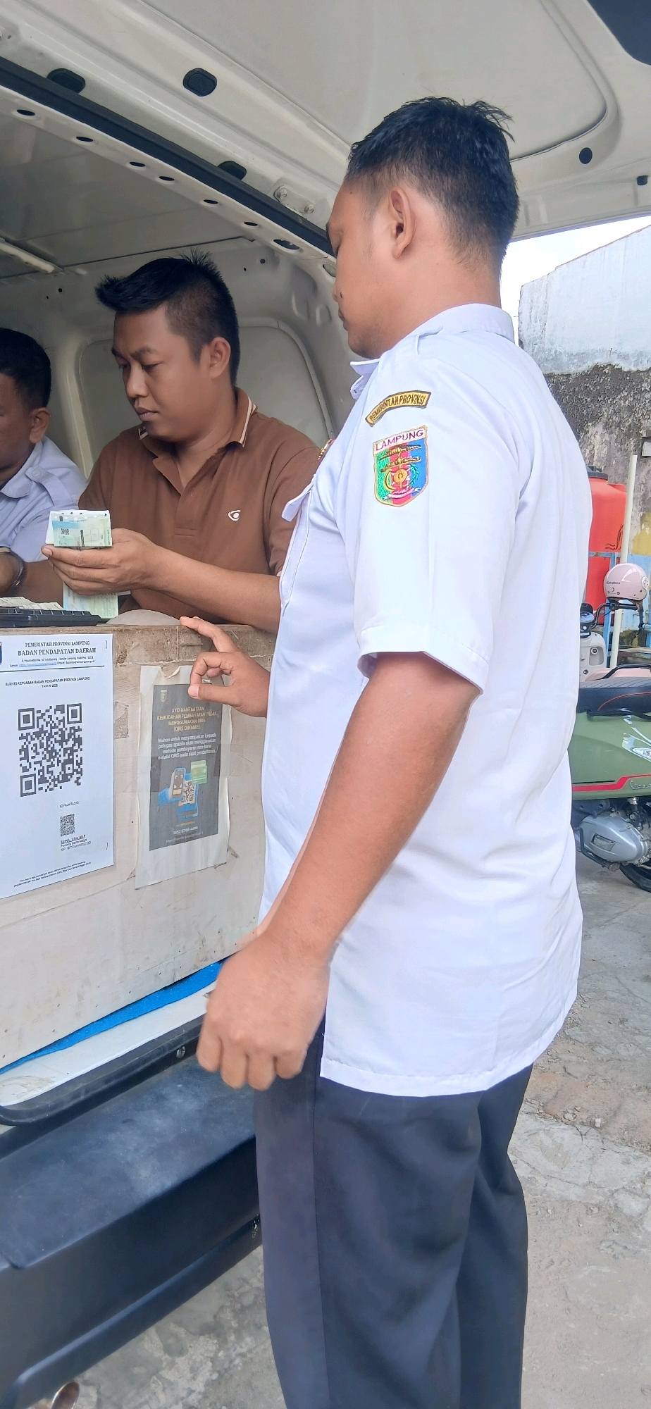 In Metro, Indonesia, a man in a white uniform with a "Lampung Provinsi" patch on the sleeve is standing next to a vehicle. He appears to be interacting with another man who is holding a wad of cash. Behind the man in the uniform, a portion of a green scooter is visible. The environment suggests an outdoor setting with a white building in the background. There are also posters on the side of the vehicle.