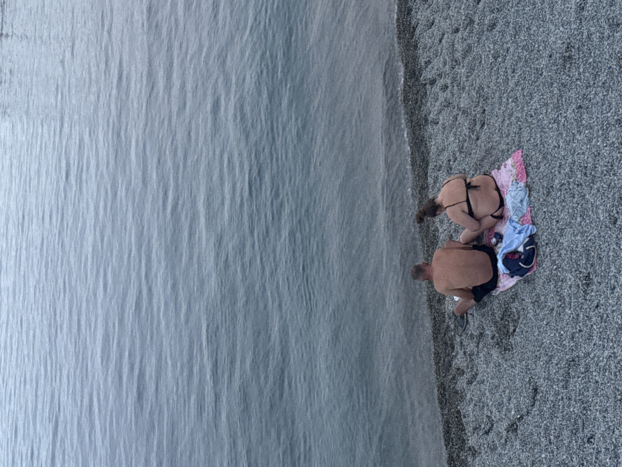 Two individuals, a man and a woman, are seated on a pebble beach in Partenit, Ukraine, facing the sea. The man, positioned to the left, is shirtless and wearing dark shorts and sandals. He is seated with his legs extended. The woman, to his right, is wearing a black bikini. She is seated with her legs bent, facing the sea and looking at her phone. They are both seated on a pink and white towel, with a blue towel and a small item that appears to be a camera or phone placed on the towel beside them. The sea is calm with gentle ripples, and the sky is overcast, contributing to a muted color palette. The beach is covered in small, gray pebbles.