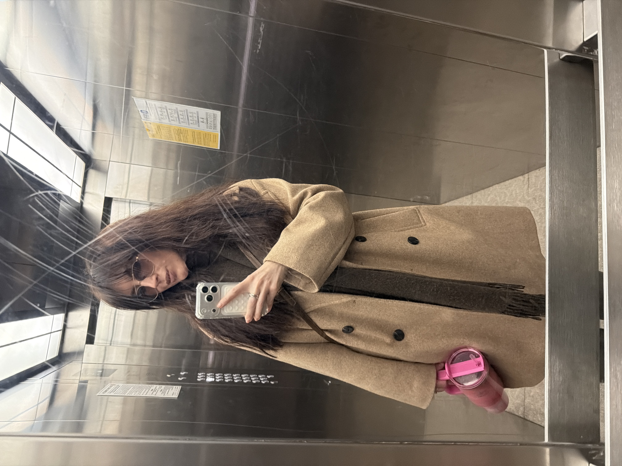 The image captures a selfie reflection of a female individual inside a brushed metallic elevator in Perm, Russia. The woman, appearing from the waist up, has long dark brown hair and wears dark aviator-style sunglasses. She is dressed in a dark turtleneck or scarf, layered under a light brown, double-breasted coat secured with dark buttons. A dark bag strap is visible diagonally across her torso. Her left hand holds a clear-cased smartphone, angled towards the reflective surface to capture the image; a ring is present on her left ring finger. Her right hand holds a translucent pink water bottle with a matching pink lid and carrying loop.
The elevator interior features scratched and reflective metallic wall panels. Reflected on the left is a control panel displaying illuminated numeric buttons, with multiple instances of "999" visible. On the right, two rectangular white information notices, containing Cyrillic text and numerical sequences, are affixed to the wall. Identifiable text fragments include "ТЕЛЕФОН" (Telephone), "8.1.1.2," "800096," "ДОМОФОН" (Intercom), and the partial company name "ООО СОЛОН," indicating the Russian context. An overhead rectangular light fixture is also reflected in the ceiling. The floor, partially visible at the bottom, appears to be light-colored.