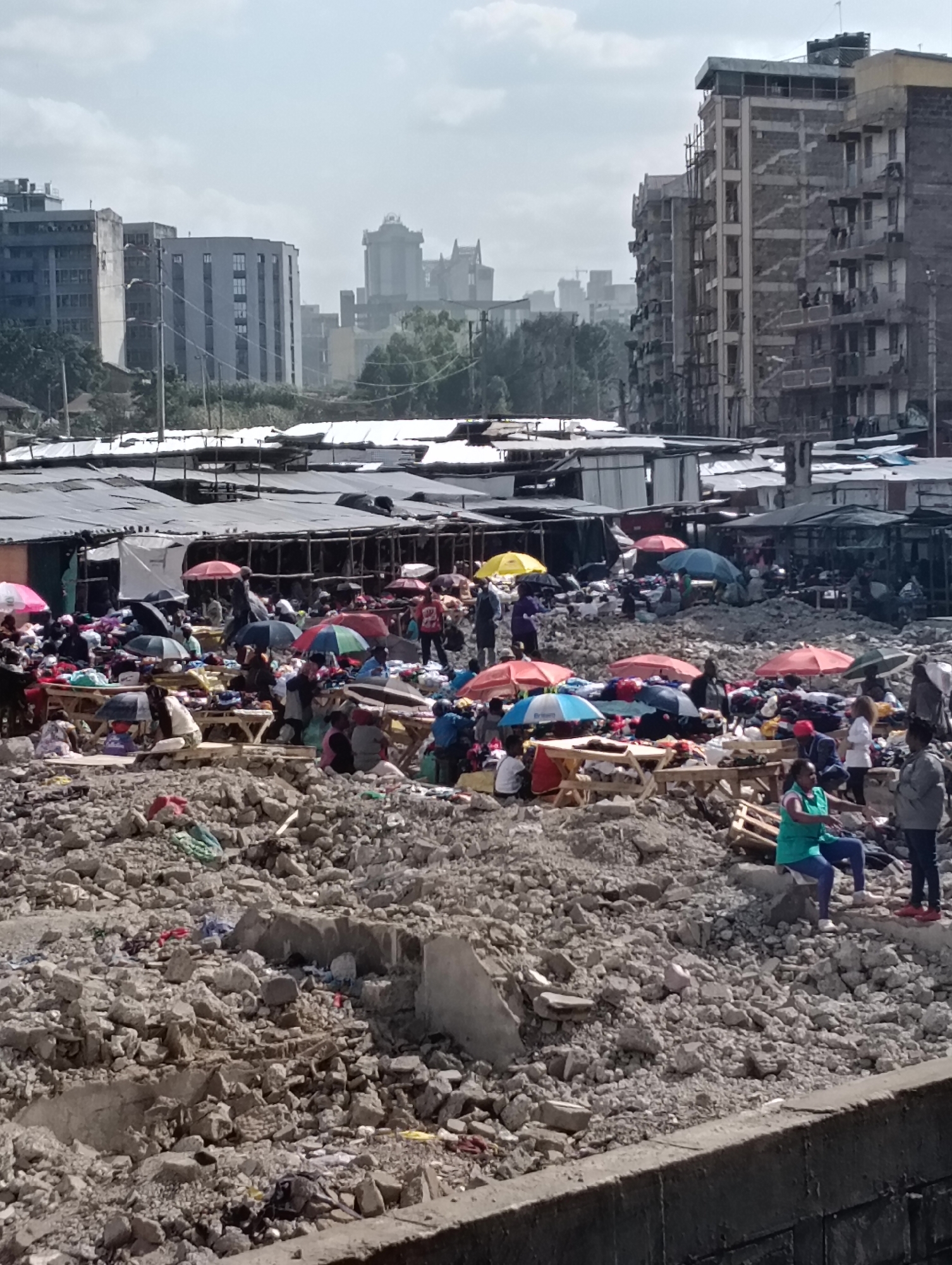 The image depicts a busy outdoor market scene in Nairobi, Kenya, characterized by a chaotic arrangement of stalls and a large amount of rubble and debris. Numerous people are present, engaged in activities typical of a marketplace, such as browsing goods, selling items, and conversing. The stalls are covered with corrugated metal roofs and tarpaulins, providing shade from the sun. A variety of goods are displayed, including clothing and textiles, laid out on tables or directly on the ground. Umbrellas of various colors are scattered throughout the market, some protecting vendors and their wares, others carried by shoppers.
The ground is covered with significant amounts of broken concrete, stones, and other construction debris, suggesting the area might be under development or has recently undergone demolition. This rough terrain adds to the visual disorder of the scene. In the background, urban buildings of varying architectural styles are visible, including apartment blocks and taller structures, indicating the market is situated within a city.
The time of day appears to be daytime, likely mid-morning to afternoon, given the brightness and the shadows cast by the stalls and umbrellas. The sky is partly cloudy, suggesting fair weather. The overall atmosphere of the market is one of bustling activity, with a sense of everyday commerce taking place amidst challenging environmental conditions. No visible text is discernable in the image.