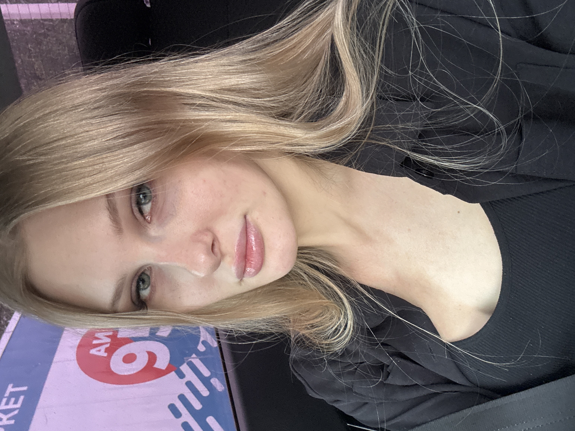 A vertically oriented, close-up shot depicts a fair-skinned person with long, blonde hair, positioned indoors with a black collared shirt or jacket. The person gazes toward the viewer with light-colored eyes, and the individual's face is slightly tilted. The background includes a glimpse of blue and pink signage with the word "KET" partially visible, along with the number "6" in a red circle. The location is identified as Perm, Russia. Additional details visible are the person's lips with a glossy appearance and sporadic freckles on the face and neck. A black seat belt strap is visible in the lower right corner. The angle suggests the photo was taken from a slightly elevated position.