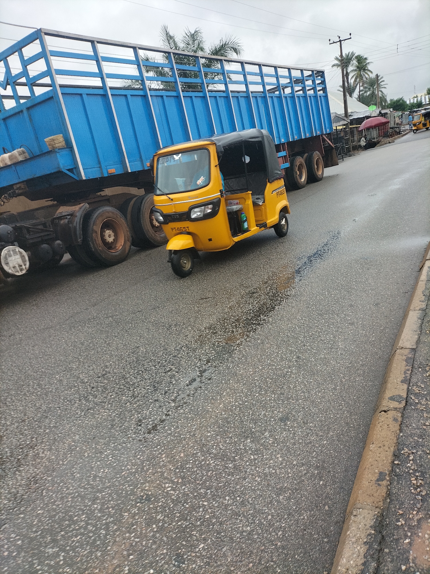 The image depicts a street scene in Jalingo, Nigeria, likely on a cloudy day. A large blue truck with a metal cage trailer is parked or moving on the right side of the road. In the foreground, a yellow auto-rickshaw, also known as a "Keke Napep" or "Marwa," is prominently featured. The auto-rickshaw has a covered passenger area and a single wheel in the front. The road surface is asphalt with visible tire marks and some wet patches, suggesting recent rain or a damp environment. Along the right side of the road, there is a raised curb and what appear to be small roadside stalls or businesses with awnings and some figures of people in the distance. Palm trees are visible in the background, contributing to a tropical or sub-tropical setting. The overall atmosphere suggests a busy urban environment with commercial activity.