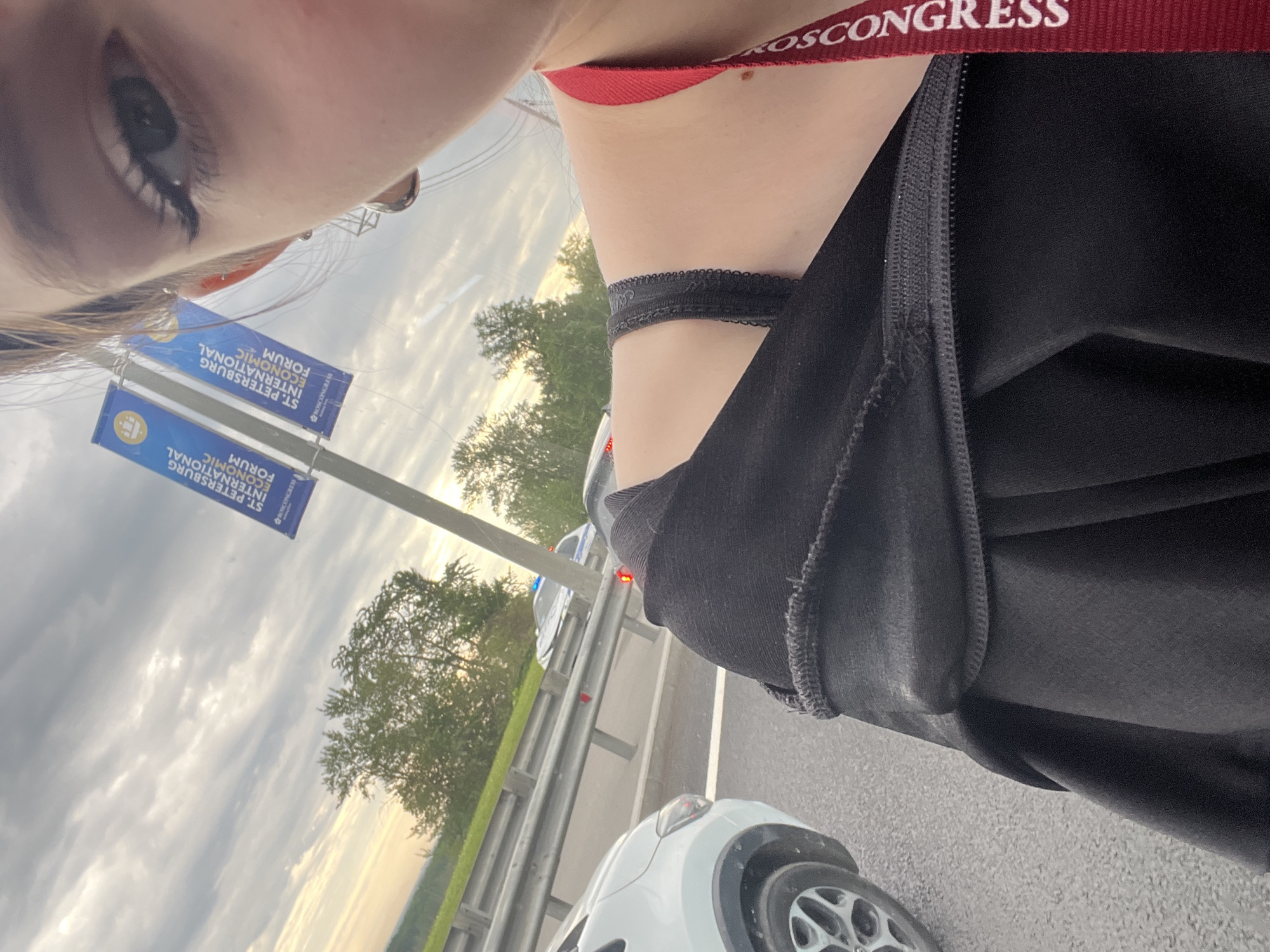 The image captures a unique, upward-angled selfie-style perspective, focusing on parts of a person and an outdoor scene. In the upper left, a close-up of a person's left eye, featuring prominent eyelashes and eyeliner, is visible. The person's neck and shoulder are seen towards the center, where a black lace choker is worn. A red lanyard, partially visible around the neck, bears the white text "ROSCONGRESS". The individual is dressed in a black garment, with a zippered detail visible on the side.
The background unfolds an outdoor setting, likely from a vehicle or roadside. The sky is overcast with soft, warm light peeking through, suggesting either late afternoon or early evening. A green, tree-lined landscape extends below the horizon. On the road, a white car, possibly an SUV, is partially visible in the foreground, and further back, another vehicle with blue and red flashing lights, indicative of an emergency or police car, is seen beyond a metal guardrail.
Two blue banners are prominently displayed on a pole in the mid-ground. The legible text on these banners reads "ST. PETERSBURG INTERNATIONAL ECONOMIC FORUM," along with a discernible logo. The combined elements point to a location near a major road or highway, likely associated with the St. Petersburg International Economic Forum, under a cloudy yet warmly lit sky.