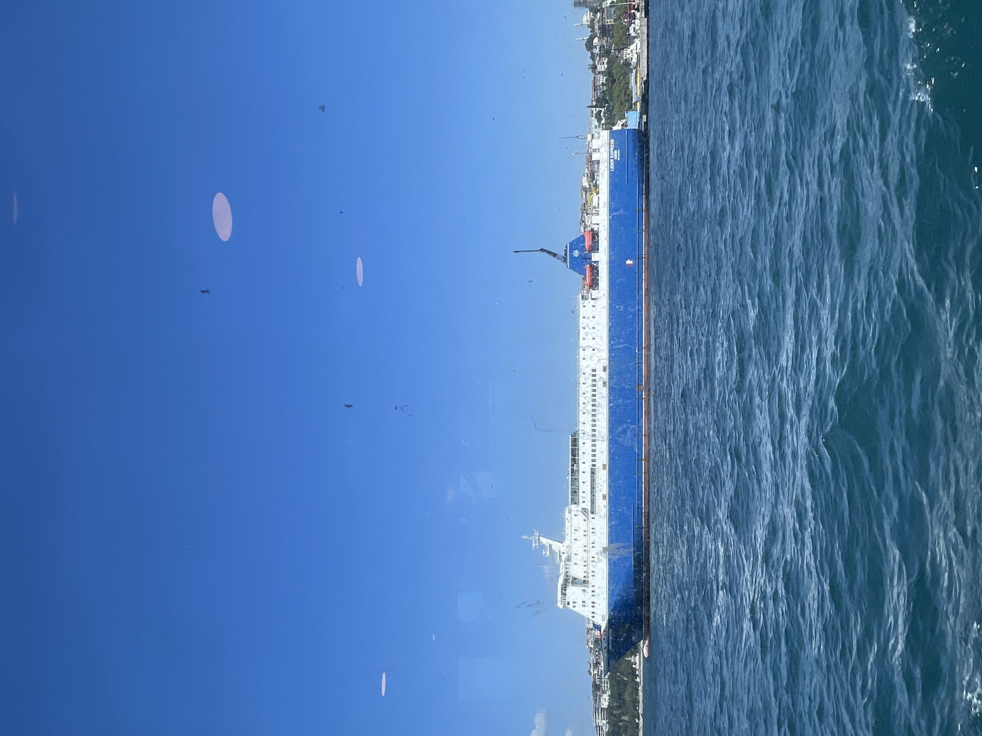 A large blue and white ferry, named "LIDER EXPRESS", is docked in the Bosphorus strait in Üsküdar, Turkey. The ferry is seen from the side, with its hull painted a vibrant blue and its superstructure a clean white. The water is a deep blue, with moderate waves. In the background, the Üsküdar shoreline is visible, featuring a mix of buildings and greenery, suggesting a populated urban area. The sky is clear and a bright blue, with a few white clouds scattered across it. The image appears to be taken from another vessel on the water.