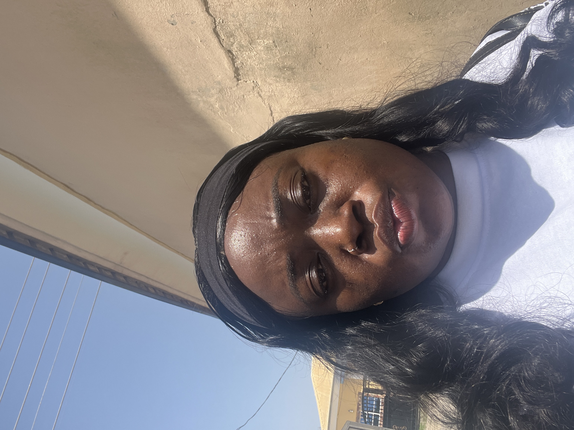 A medium close-up shot, taken outdoors during daylight, depicts a dark-skinned woman with black hair and a black headband. She is wearing a white shirt. The sun appears to be shining directly onto her face. Behind her are light-colored structures, possibly buildings, and a patch of blue sky. The location is possibly within Abuja, Nigeria. No specific interactions or actions can be discerned.
