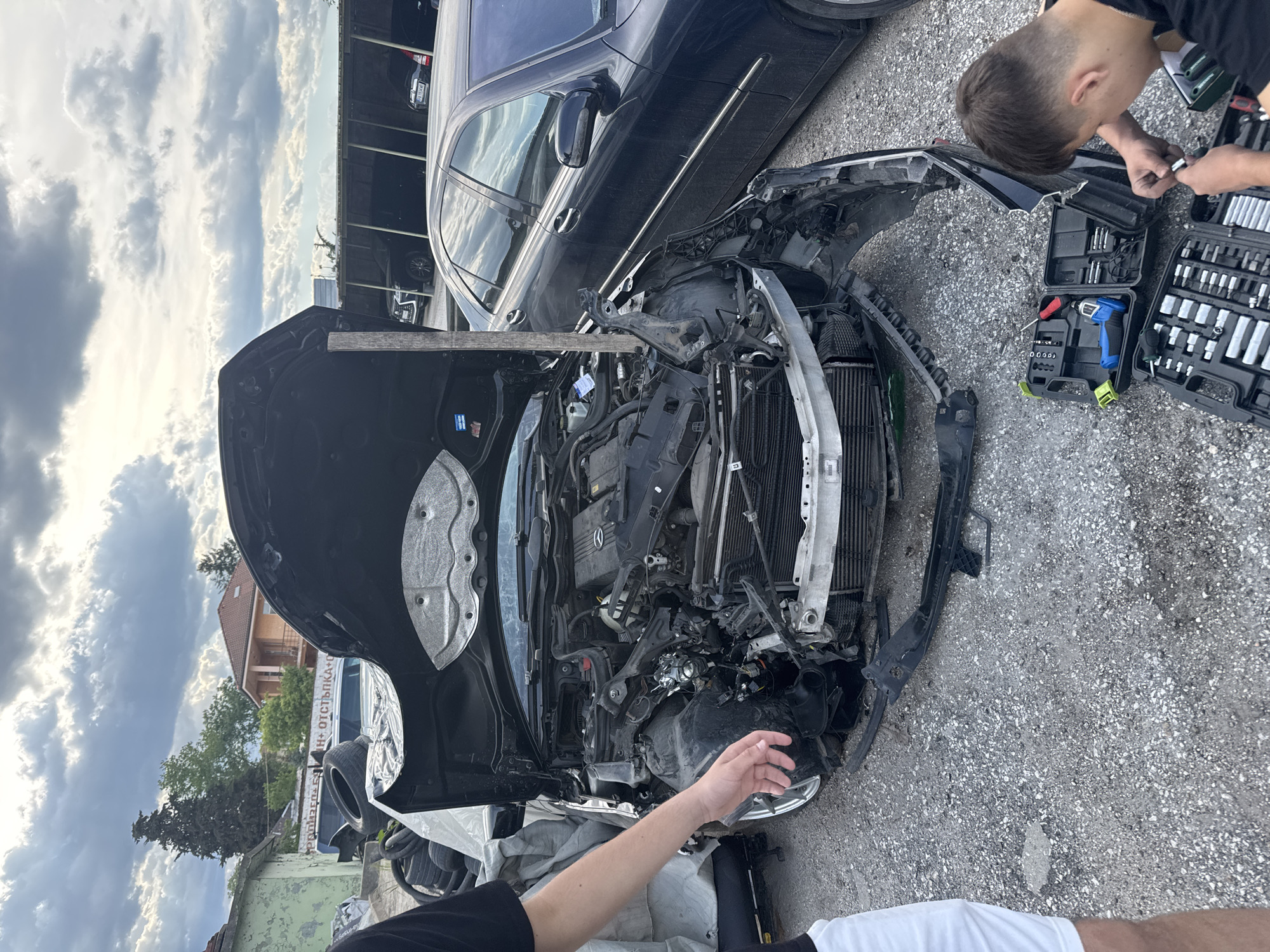 A car with severe front-end damage is in a parking lot in Pazardzhik, Bulgaria. The car’s hood is open and supported by a wooden beam. The front of the car is completely destroyed, exposing the engine and radiator. A man is working on the car's front right. The environment is an outdoor parking lot with other cars parked nearby. A second man stands near the car and appears to be working on it using a set of tools. A building is visible in the background. The sky is overcast with storm clouds.