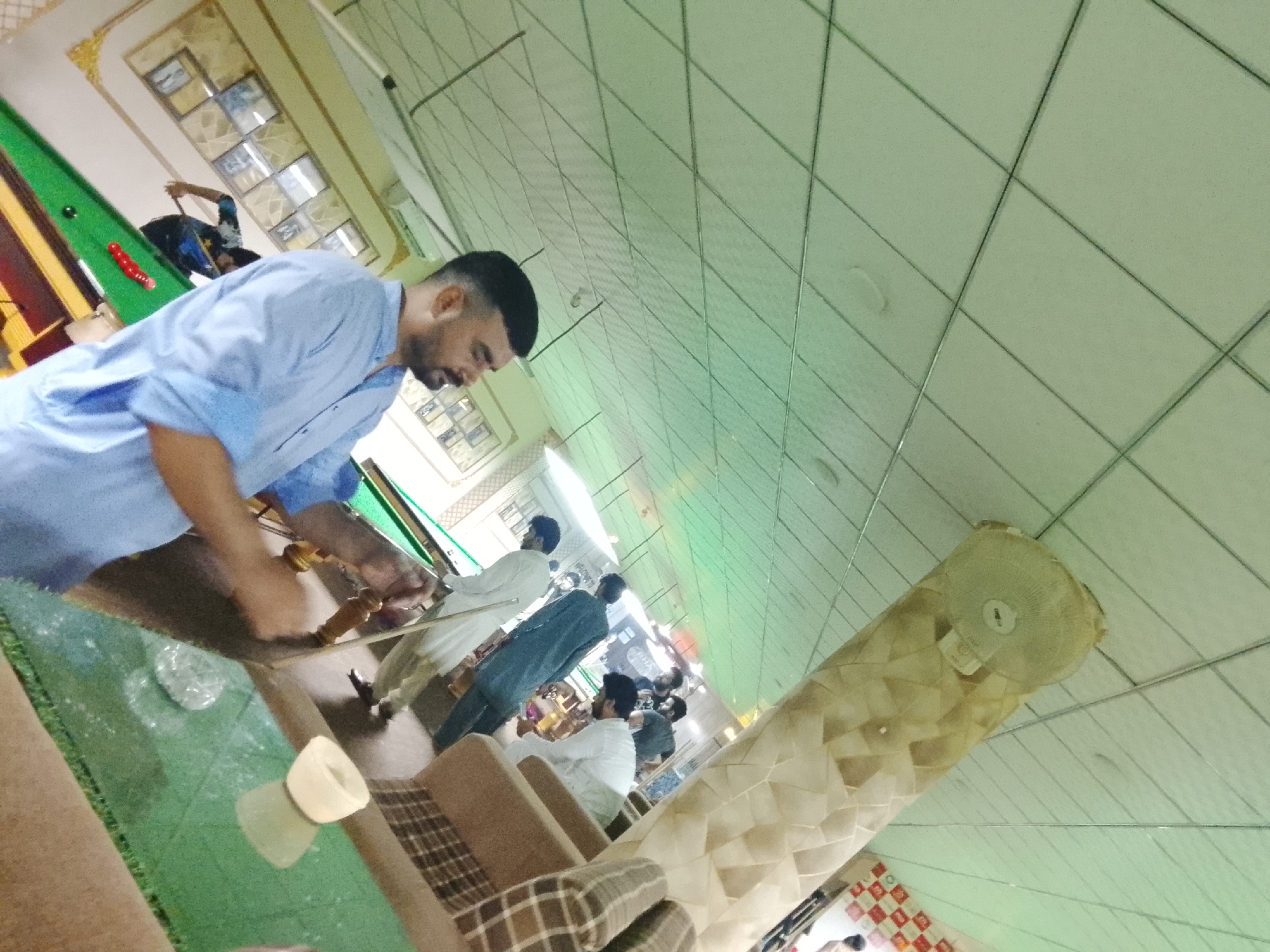 The image depicts an indoor recreational setting, likely a snooker or pool hall, filled with several men. The scene is captured at an oblique angle.
In the foreground, a man with a dark beard and short hair, wearing a light blue long-sleeved shirt, leans forward over a table, holding a snooker cue. He appears focused on the table's surface, which reflects parts of the room. A clear plastic bottle is also visible on this table.
Behind him and to the left, a green snooker table is visible with red and a black ball. Another man, wearing a dark shirt, is actively playing on this table with a cue. Further into the background, other individuals are present. One man in a white traditional outfit (possibly shalwar kameez) stands by another snooker table, while a man in a dark blue garment also observes or plays. More men are seated on patterned sofas or standing further in the background.
The setting features light-colored ceiling tiles arranged in a grid pattern. A large, textured, decorative column-like structure with a fan mounted on it stands prominently in the mid-ground. The lighting appears to be artificial, typical of an indoor entertainment venue, making the time of day undeterminable. The atmosphere seems relaxed, with people engaged in snooker or socializing. No prominent text is clearly legible.