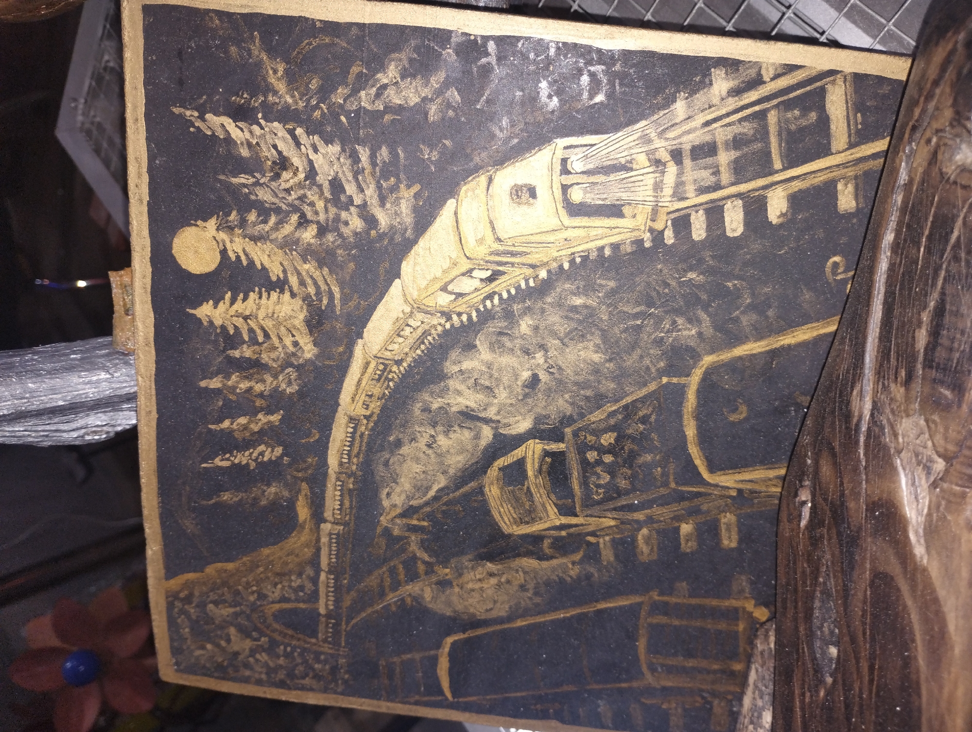 The image is a painting on a dark surface, possibly a canvas or board, with gold paint used for the artwork. The painting depicts a train on a track at night. In the foreground, a train with its headlights on is moving on a track. The track curves to the left and goes under a tunnel. The train itself is long, with multiple carriages. In the background, there are stylized pine trees and a full moon in the night sky. The overall style of the painting is somewhat primitive and abstract. There is no indication of the city of Perea, Greece, within the image itself. The painting is positioned on a rustic wooden surface.