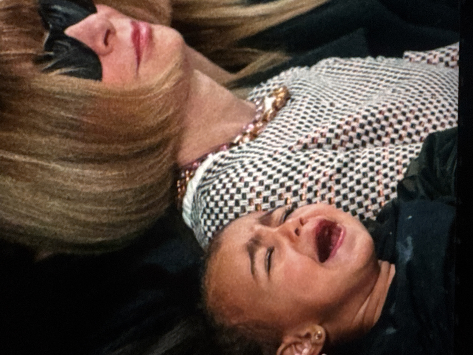 The image shows a close-up of a young child crying next to a woman wearing sunglasses. The child has their eyes closed and mouth open in distress, with a small gold earring visible in their left ear. The woman has blonde hair and is wearing a patterned top with a beaded necklace. The background is dark and out of focus. The image does not contain any elements that suggest a location in Kuta Selatan, Indonesia.