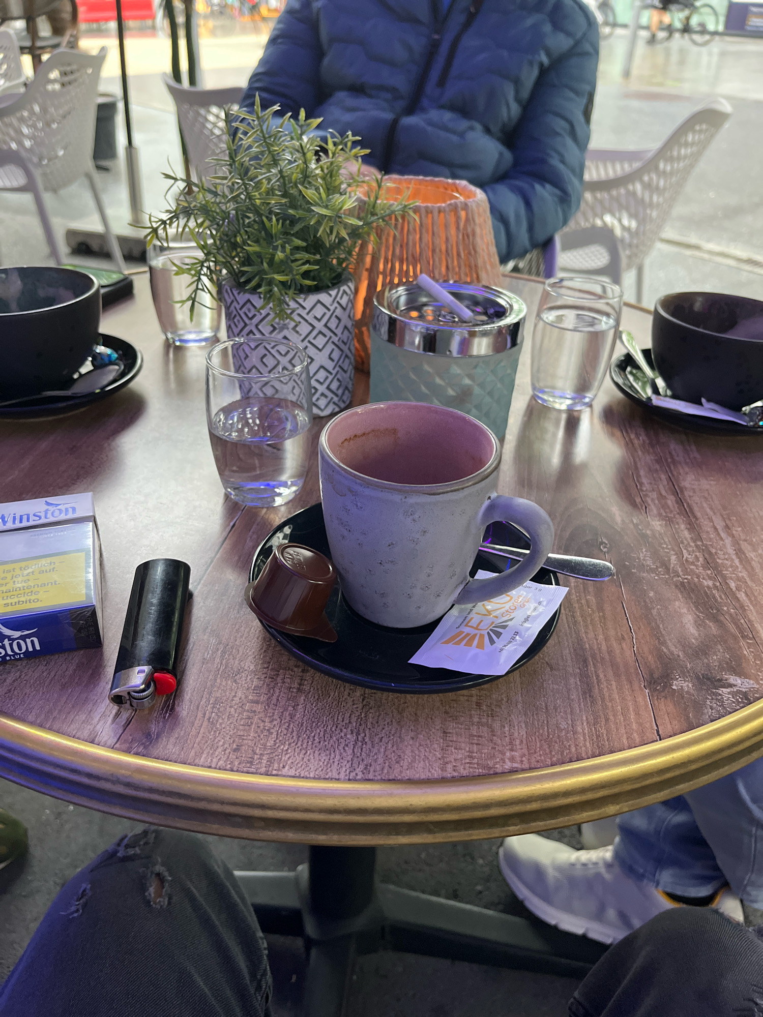 The image depicts an outdoor seating area at a cafe with a wooden table. On the table are two dark-colored mugs, one with a pink interior, a pack of Winston cigarettes, a black lighter with a red flame button, an ashtray with a cigarette, and two glasses of water. A small decorative plant in a patterned pot sits near the ashtray, and a small brown dessert is placed on a saucer with a sugar packet and a spoon next to the mug with the pink interior. In the background, blurred figures of people are visible, and one person is wearing a blue puffer jacket. Another person's legs are visible in the foreground, wearing ripped jeans and white sneakers. The overall setting appears to be a casual outdoor cafe environment. The location is identified as Buchs (SG), Switzerland.