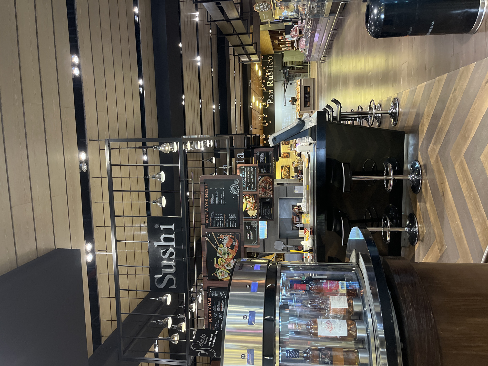 The image shows the interior of a store or market in San Bernardino Tlaxcalancingo, Mexico. The scene is focused on the food service area, displaying offerings like sushi and pan rustico (rustic bread). A black sign with white lettering spells out "Sushi." Menus advertise "POKE & YAKIMESHI" and "SUSHI Y MÁS". There's a refrigerated display case containing bottles, possibly wine, along with a service counter equipped with bar stools. Above the food service areas are signs for "Pan Rustico" and "QUESOS DEL MUNDO". The interior design includes wooden wall panels and a metal framework, with lighting fixtures hanging throughout.