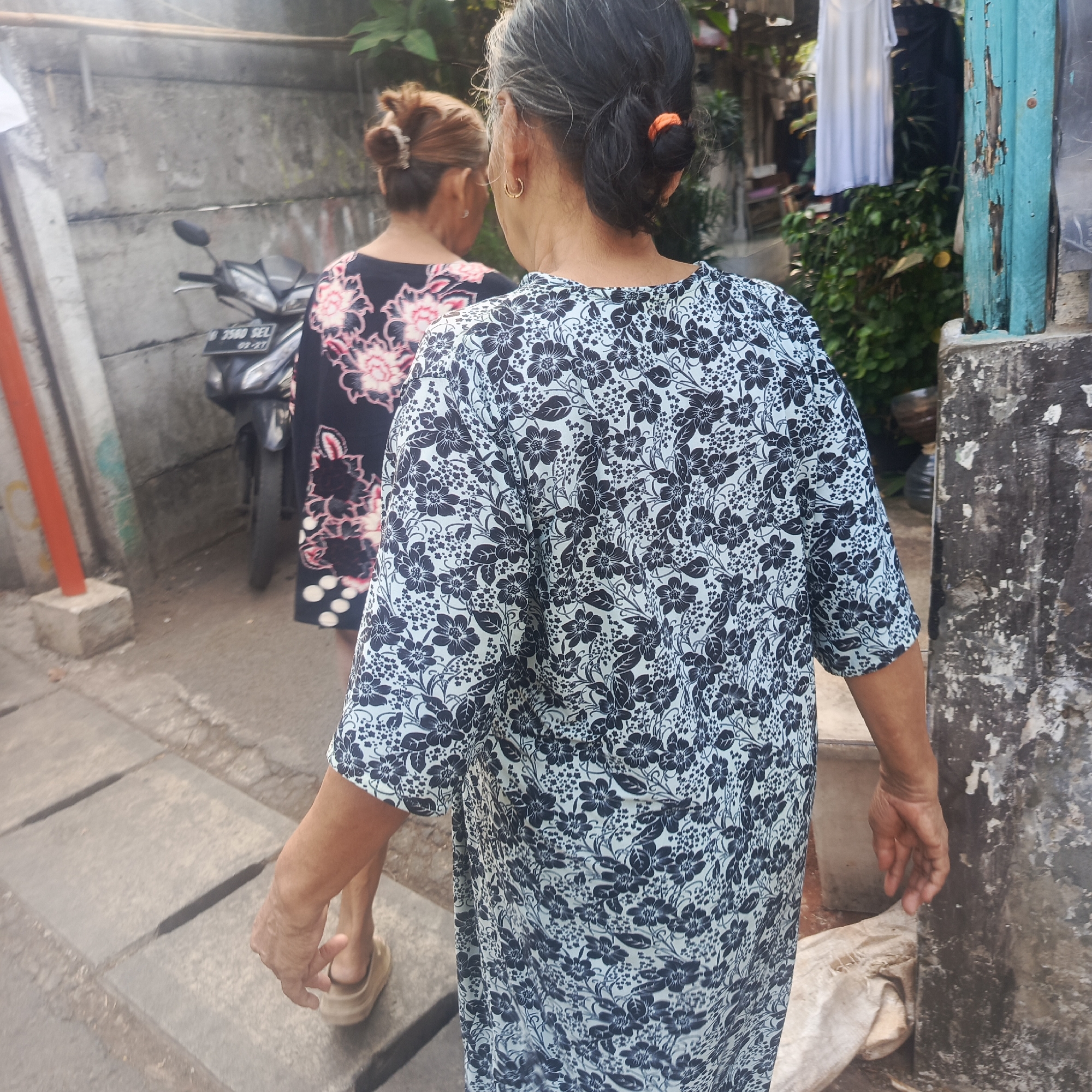 Two individuals, viewed from behind, are walking down a paved alleyway. The person in the foreground wears a light blue dress with a dark blue floral pattern. The person behind them wears a dark dress with a pink floral pattern. A motorcycle is parked to the left of the alleyway, and various structures and vegetation are visible in the background. The scene appears to be in an urban environment, possibly within the city of South Jakarta, Indonesia. The lighting suggests it is daytime.