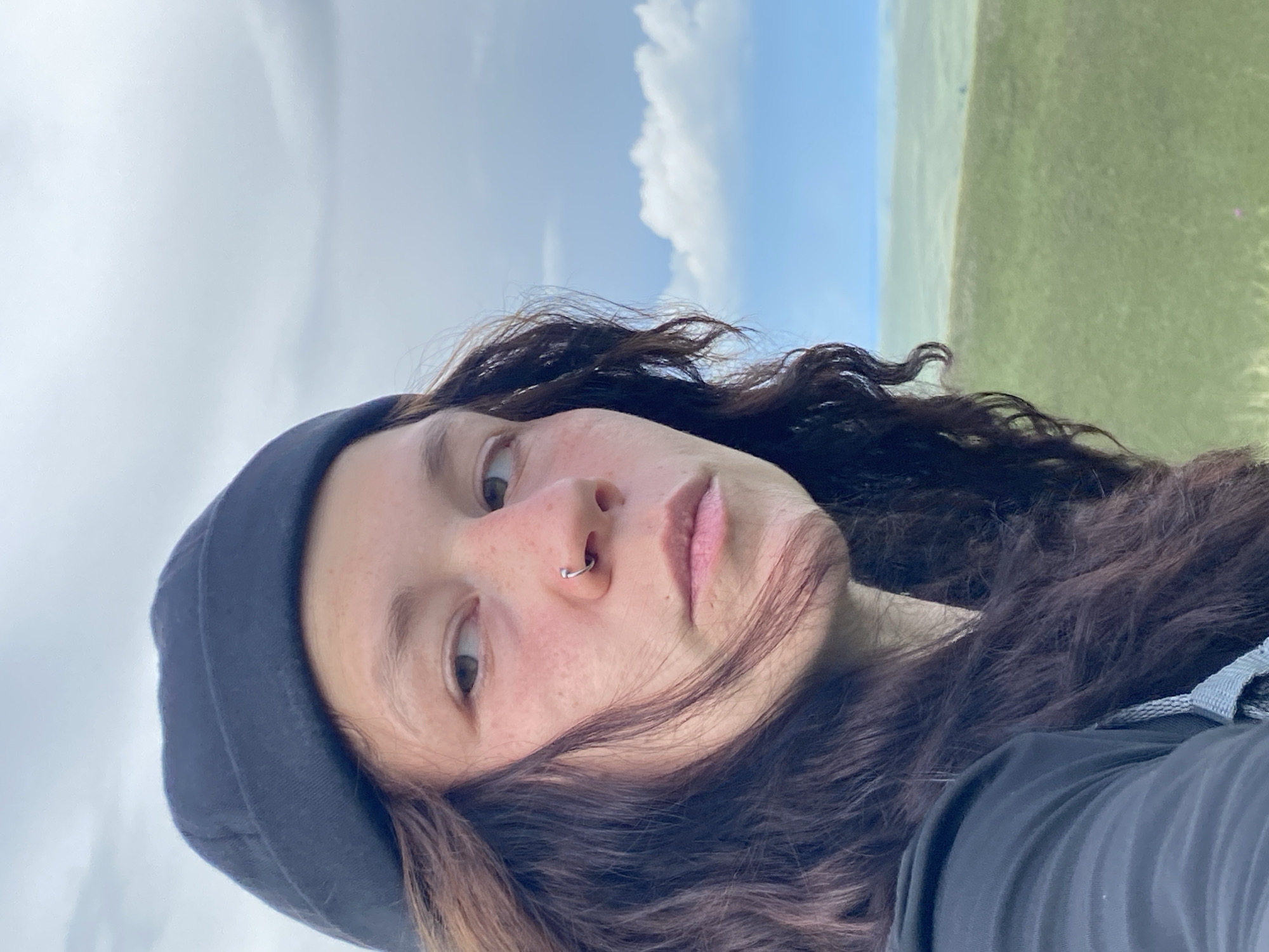 A close-up, head-and-shoulders portrait of a young person wearing a black beanie, their face turned to the right, looking off-camera. They have long, dark, wavy hair, prominent cheekbones, and a nose ring. Their skin is fair with a smattering of freckles. The background shows a landscape with rolling green hills under a cloudy sky, typical of the Verkhneozernoe, Russia area. The lighting suggests it is daytime, likely overcast with some brighter areas in the sky.