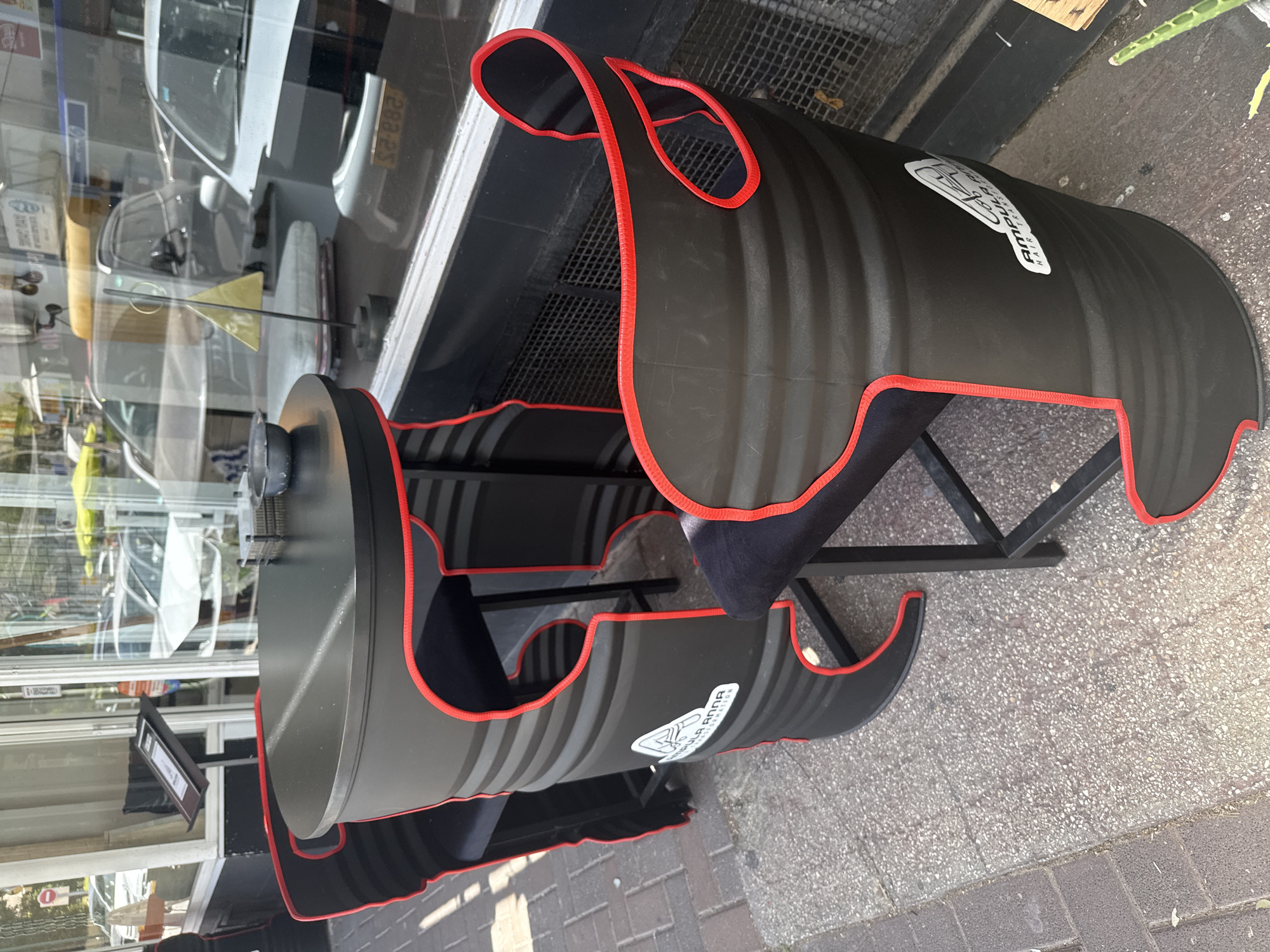 A black, barrel-shaped chair with red trim sits on a sidewalk next to a glass storefront. The chair has cut-outs and what appears to be a cushion for seating. The storefront reflection shows a white car and other objects. The image was taken in Raanana, Israel.