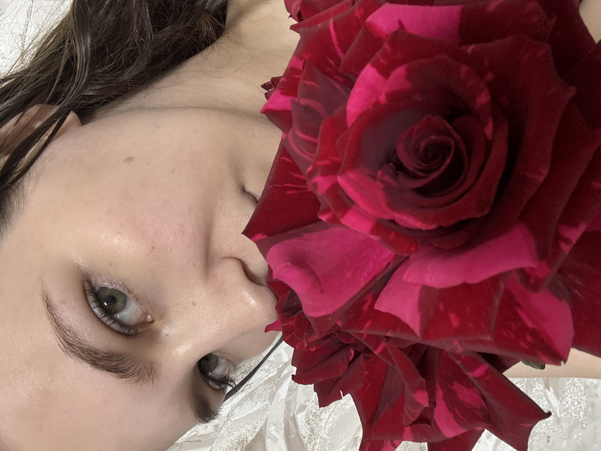 A close-up, high-angle portrait features the upper half of an individual's face partially obscured by a bouquet of variegated red and fuchsia roses. The subject, appearing female, has dark brown, wavy hair, seemingly damp, visible on the right side of the frame. Her hazel or green eyes are open, looking directly at the viewer, with distinct eyebrows. Fair skin with some natural texture is visible on the forehead, cheek, and nose, where a slight sheen is present. A small, dark brown mole is located on the right cheek (viewer's left). The lower portion of the face is covered by numerous rose blooms, primarily a vibrant, streaky blend of deep red and fuchsia, with one tightly coiled rose head prominent in the lower right. A small portion of the subject's neck and left shoulder is visible above and behind the roses. The background consists of a light-colored, possibly off-white, textured fabric, resembling bedding or a pillow. The lighting is soft and even. The image provides no discernible visual cues regarding a specific geographical location, including the city of Chaikovsky, Russia.