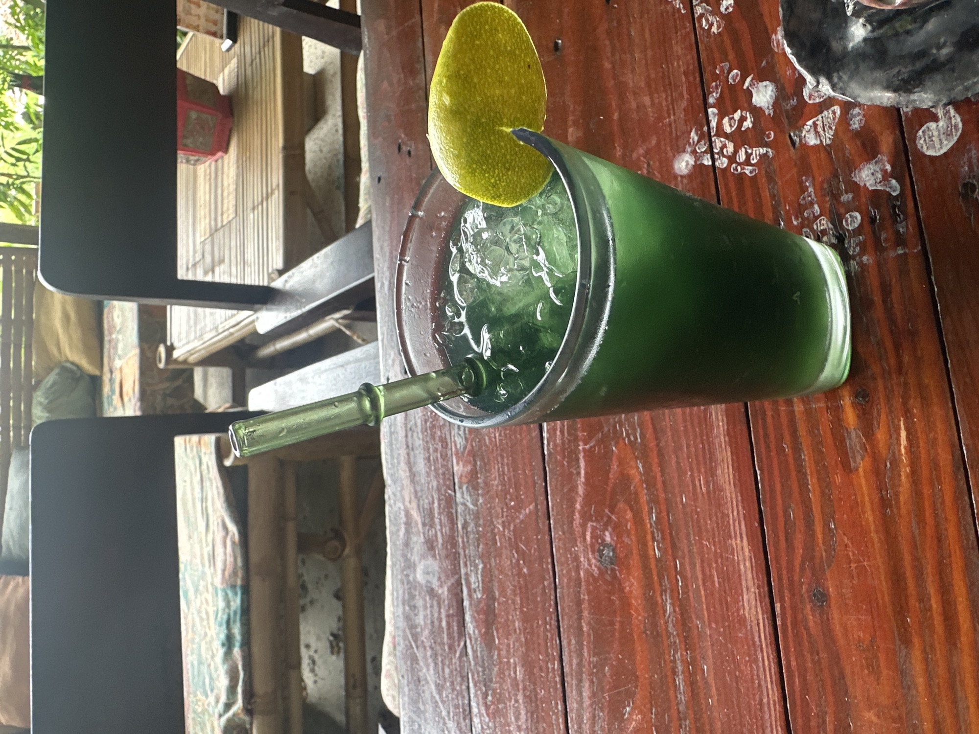 A green beverage with ice and a slice of lime sits in a glass on a wooden table. A glass straw extends from the drink. In the background, there are dark chairs, bamboo furniture, and foliage. The location is a cafe or restaurant in Koh Phangan, Thailand.