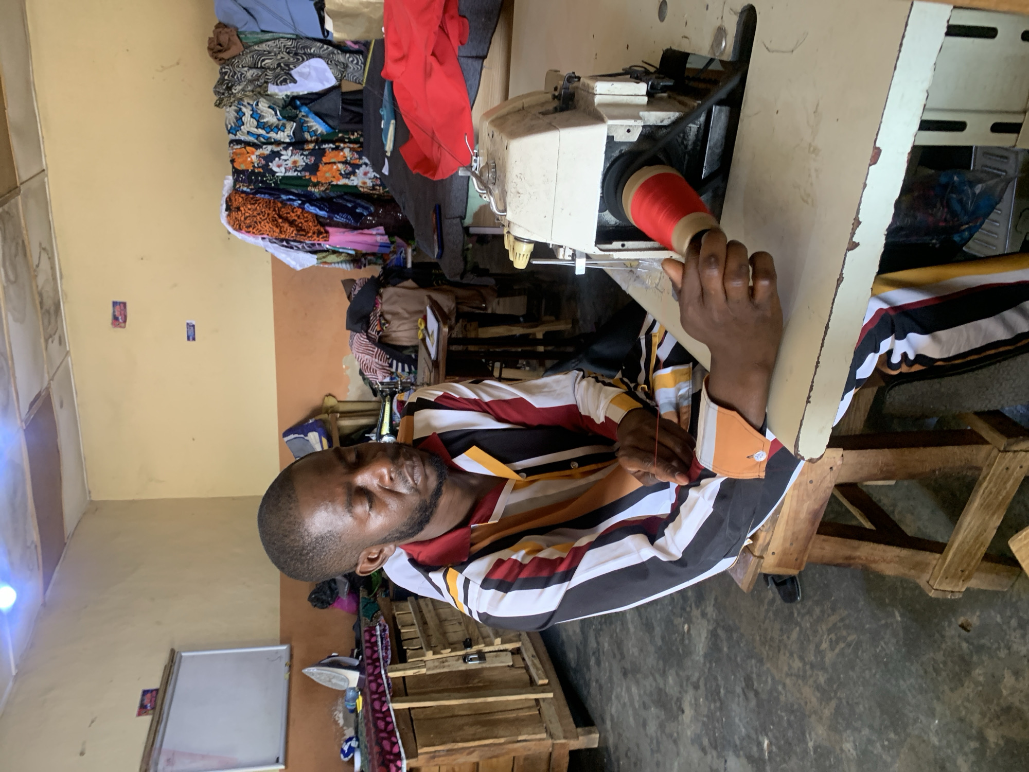 The image shows a man in a striped shirt, seated and threading a sewing machine with a large spool of red thread. The sewing machine is on a white table. The man's skin tone is dark. Behind the sewing machine and to the left are piles of folded and stacked fabrics in various colors and patterns. A wooden structure, possibly a stool or table, is under the white table. An iron sits on a wooden dresser. The background is a peach-colored wall, with light coming from the left. The location is presumed to be a tailor's shop in Jos, Nigeria.