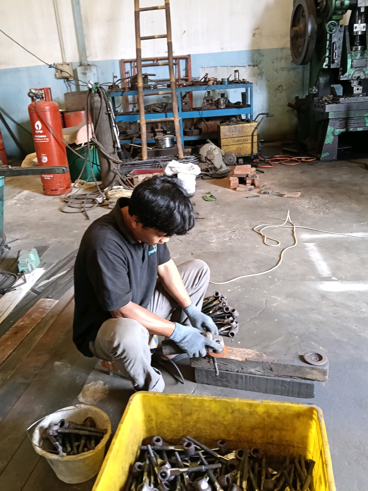In a workshop located in Belimbing, Indonesia, a man is seen working. He is crouched on the floor, wearing a black polo shirt and grey pants, with blue gloves on his hands. He is actively grinding a metal object with a small grinder, which is placed on a wooden block. In front of him, a pile of metal pieces is gathered, and to his right, a yellow plastic bin overflows with similar metal parts. The workshop environment is evident from the presence of tools, machinery, and various industrial materials scattered around the concrete floor, including a red gas cylinder, shelving units, and electrical wiring. The lighting suggests an indoor setting with overhead illumination.