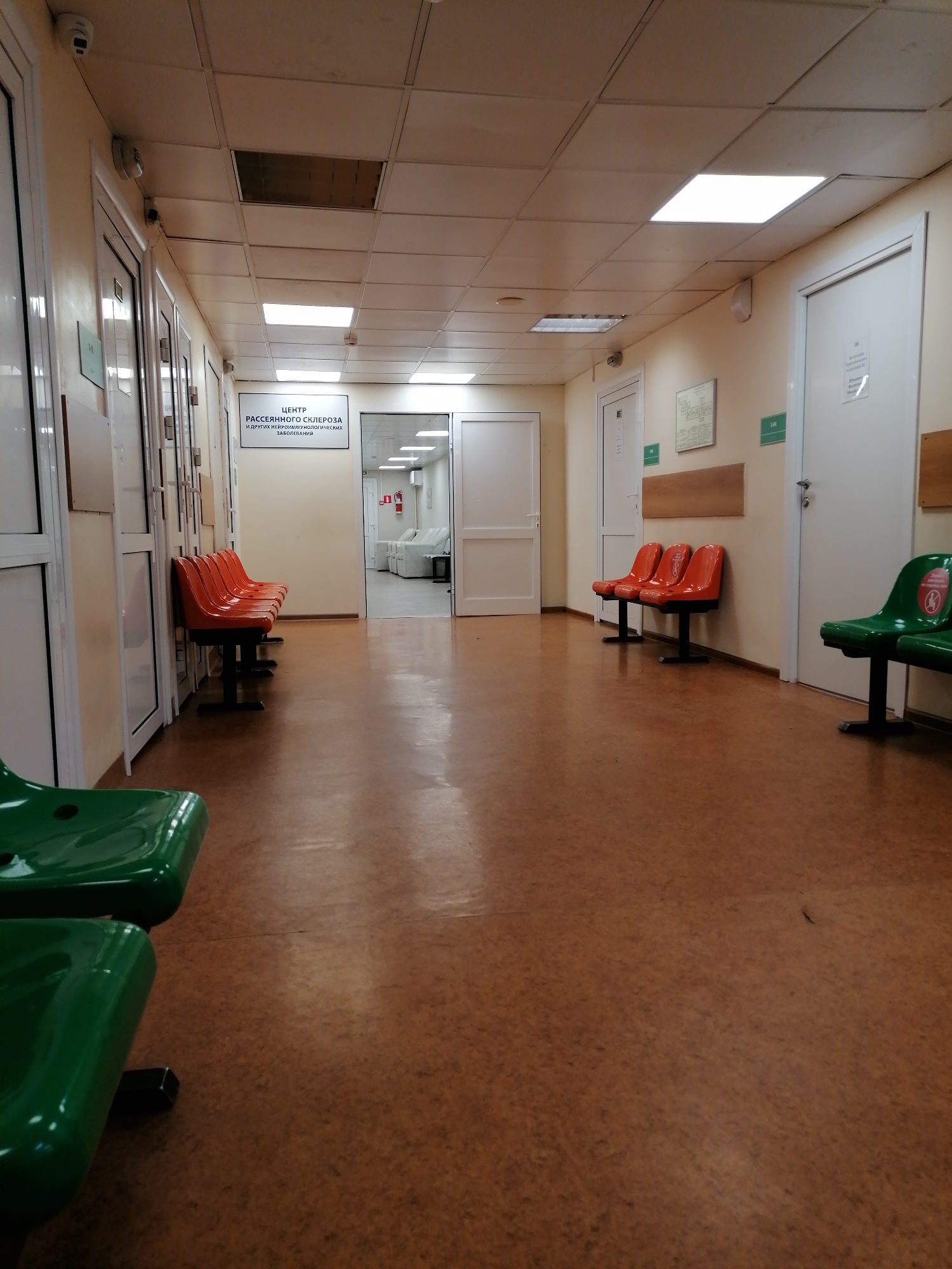 An eye-level, slightly low-angle shot captures an empty, well-lit hospital or clinic hallway. The long corridor features light-colored walls, possibly cream or light yellow, and a reddish-brown floor, likely linoleum, that reflects some overhead light. The ceiling consists of white acoustic tiles with several rectangular fluorescent light panels, some illuminated, providing bright, artificial illumination.
Along the left side of the hallway, several white doors with glass panels and metal frames are visible, suggesting multiple rooms. In front of these doors, a row of four bright orange waiting chairs with black metal frames is positioned. Further down the hall, a large white sign is mounted on the wall. The sign, written in Cyrillic, reads "ЦЕНТР РАССЕЯННОГО СКЛЕРОЗА И ДРУГИХ НЕЙРОИММУНОЛОГИЧЕСКИХ ЗАБОЛЕВАНИЙ," which translates to "CENTER FOR MULTIPLE SCLEROSIS AND OTHER NEUROIMMUNOLOGICAL DISEASES." Beyond this sign, an open doorway reveals a brightly lit room with several white beds or examination couches.
On the right side of the hallway, more white doors are present, some with small, unreadable posters. Two groups of orange waiting chairs are visible against this wall. In the immediate foreground on the right, two bright green waiting chairs with black frames are positioned, one featuring a red circular "do not sit" or social distancing sticker. Small surveillance cameras are mounted near the ceiling at various points.
The scene is devoid of people, contributing to a quiet, somewhat sterile atmosphere typical of a medical facility. The time of day is not discernible due to the exclusive use of artificial lighting.