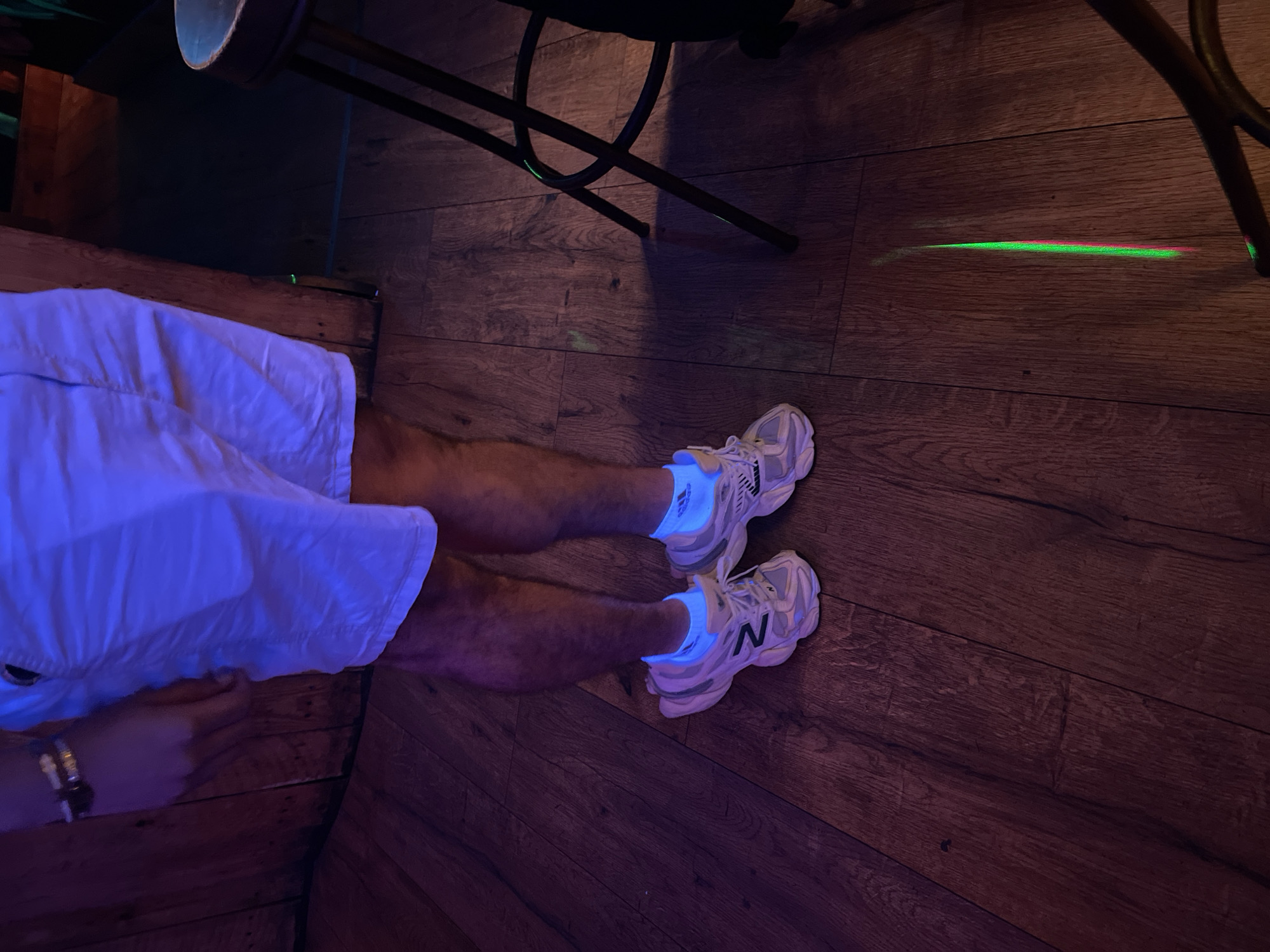 The image shows a close-up of a person's legs and feet, standing on a wooden floor. The person is wearing white shorts, light-colored athletic shoes with blue socks, and a bracelet on their left wrist. The environment appears to be dimly lit, with purple and blue lighting, and there is a bar stool visible to the right of the frame. A green and pink light streak is visible on the floor. The image does not provide any specific details that would allow for identification of the location as Barcelona, Spain.