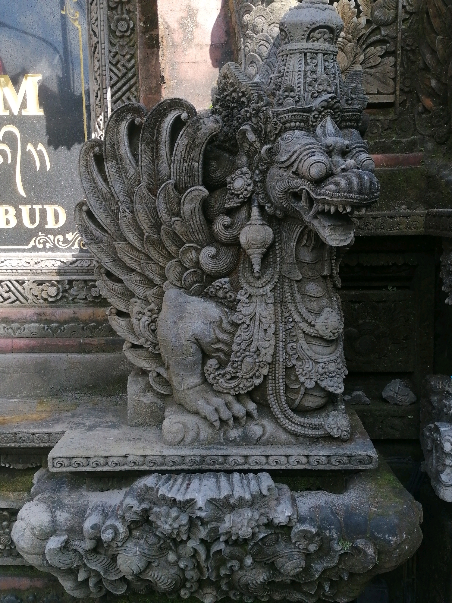 This image shows a stone statue of a mythical creature, likely a dragon or a similar guardian spirit, in front of a stone building with ornate carvings. The statue is predominantly gray and weathered, with intricate details in its scales, wings, and facial features. The creature has a fierce expression, with its mouth open revealing sharp teeth. It is adorned with detailed necklaces and a crown-like headdress. Behind the statue, the building features a dark stone facade with carved patterns and some moss growth, suggesting an outdoor setting. To the left of the statue, a black stone monument with golden lettering is visible, with the partial words "M," "BUD," and some stylized script. This scene is located in the Ubud District, Indonesia, an area known for its rich cultural heritage and traditional art. The lighting suggests it is daytime, with some shadows indicating the direction of the sun. No people are present in the image.