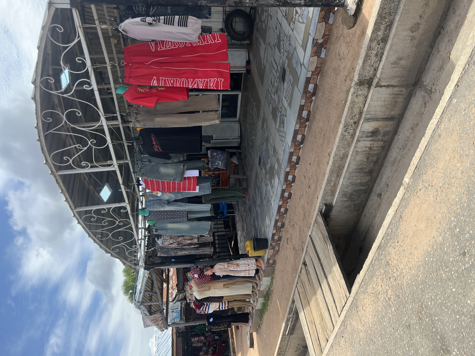 This detailed description depicts a street scene in Jalingo, Nigeria, focusing on a clothing stall or shop.
The scene is an outdoor setting under a partly cloudy sky, with patches of bright blue interspersed with white and light gray clouds. In the foreground, an asphalt road surface, lightly dusted with sand or dirt, occupies the bottom left. Adjacent to the road is a concrete drainage ditch or culvert, which extends across the lower portion of the image. A makeshift wooden bridge, constructed from several planks, spans a section of this ditch on the left, leading presumably to the shop's entrance.
Behind the ditch, a narrow strip of reddish-brown dirt separates the drainage system from a raised platform that serves as the shop's immediate frontage. This platform is tiled with a mosaic pattern of grey, beige, and light brown squares, and its vertical edge features a decorative border of brown and white patterned tiles.
The main subject is a clothing shop operating under a curved corrugated metal roof, supported by visible metal framework. A white, ornate wrought-iron decorative element is attached to the front edge of the roof structure. A square fluorescent light panel is visible mounted underneath the roof.
Various articles of clothing are displayed, hanging on hangers from the roof structure. These include blue jeans, a black t-shirt with "Highway" printed in white, grey trousers, red t-shirts, and prominent red track pants with white vertical lettering. The red track pants on the left display "MAMUNITY" (possibly mirrored "COMMUNITY"), while the pair on the right reads "VILLOVIAN" (possibly mirrored "ALLOVIAN"). Other items include striped shirts and various patterned garments.
A row of seven headless mannequins stands on the tiled platform to the left. These mannequins display different outfits, including dresses and pantsuits in various colors and patterns. One smaller mannequin, possibly representing a child or petite adult, wears a long floral dress. To the right of the mannequins, a yellow jerrycan and a black bucket rest on the ground.
Inside the shop, visible through the open front, a blue plastic chair, a black tire leaning against a wall, and what appears to be a broom are discernible. To the far left of the main shop, an additional, less elaborate stall with a corrugated metal roof is partially visible. Green palm fronds are visible in the background above the roofline of the structures.