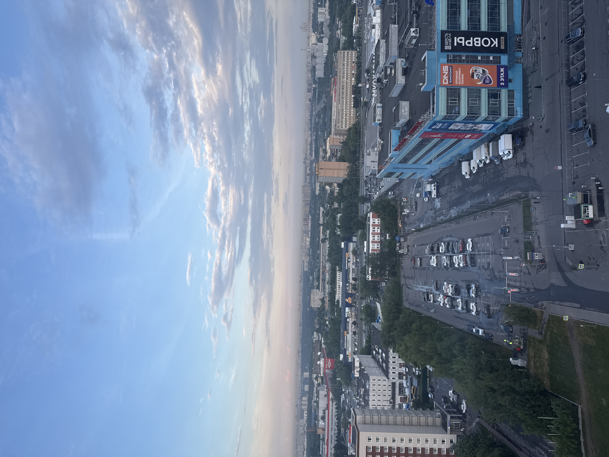 The image captures a panoramic view of a cityscape, likely Moscow, Russia, under a hazy, overcast sky with scattered clouds. In the foreground, a large, multi-story blue commercial building with prominent signage, including "DNS Технопоинт" and "КОВРЫ", dominates the right side of the frame. Adjacent to this building is a wet, paved parking lot filled with numerous cars. Further left, a multi-story residential building with a white facade and numerous windows stands beside a section of dense green trees. In the mid-ground, a sprawling urban landscape unfolds with various industrial and commercial structures, interspersed with more trees and what appear to be smaller residential buildings. A distant horizon is visible, marked by a faint line of buildings and a hazy sky. The overall atmosphere suggests an urban environment, possibly at dawn or dusk due to the diffused lighting.