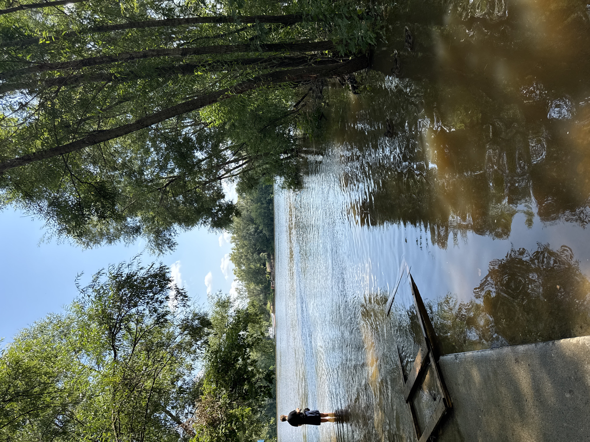 The image depicts a wide, muddy river on a sunny day. On the left bank, a person stands partially submerged in the water, facing away from the camera. They are wearing a dark t-shirt and shorts. A concrete ramp leads into the water, with a wooden frame structure floating partially submerged. The right bank is lined with dense trees. In the distance, across the river, more trees and a small white boat are visible. The water is calm with ripples. The sky is blue with scattered clouds. The scene appears to be a natural setting on the outskirts of a city, possibly Ufa, Russia, given the context.