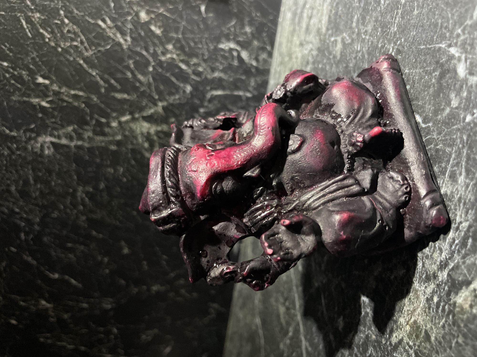 The image shows a small, intricately detailed sculpture of a deity, likely Ganesha, in a dark red and black color scheme. It is positioned against a grey, textured surface reminiscent of marble or stone. The lighting casts a dramatic shadow, emphasizing the form of the sculpture.
There is no information in the image to suggest the location is Kazan, Russia, and I cannot provide information about the city.