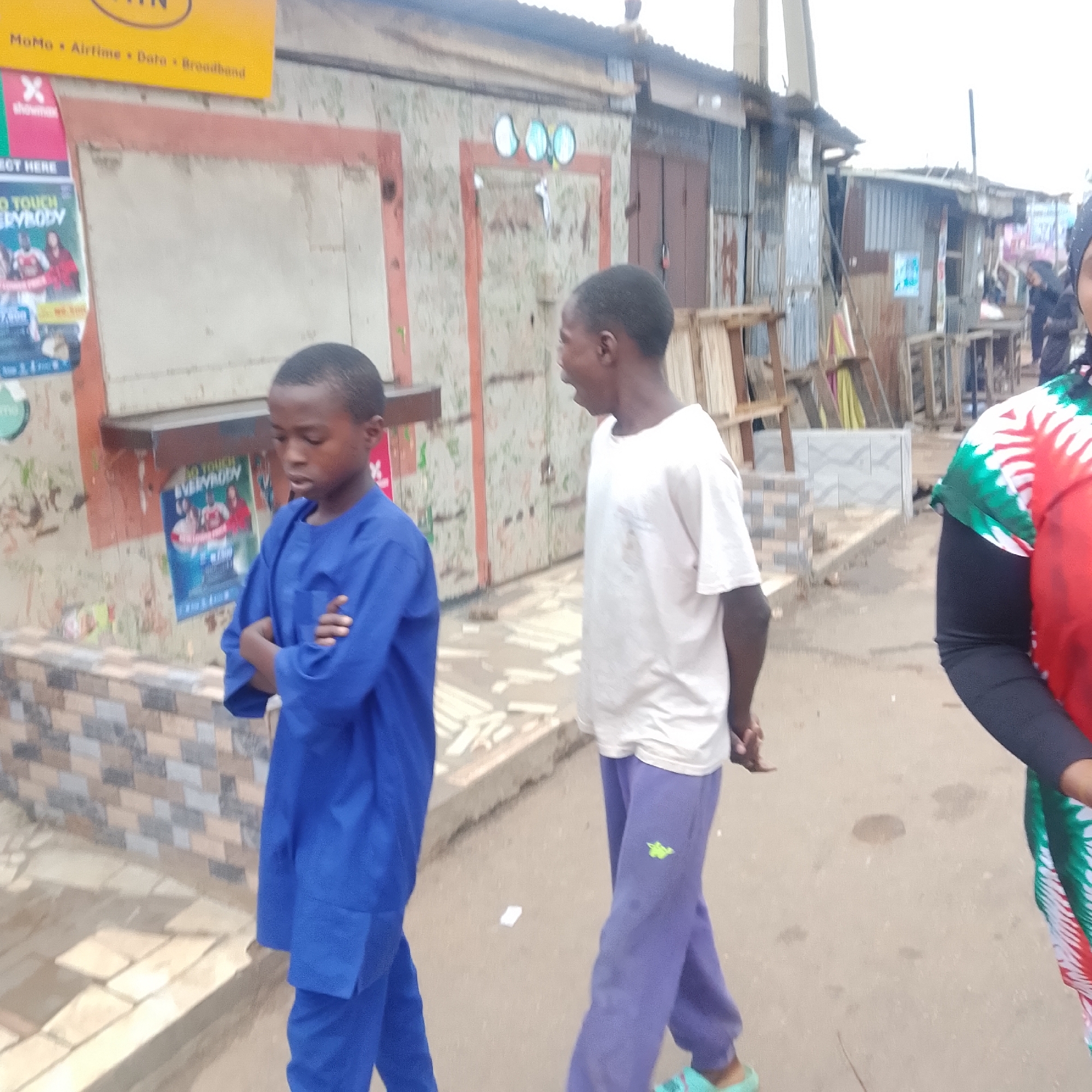 The image shows a street scene with several people and structures. In the foreground, two young boys are walking. The boy on the left is wearing a blue outfit, and the boy on the right is wearing a white t-shirt and purple pants. Further in the background, a person in a red, green, and white shirt is visible. The environment appears to be a market or a street lined with small shops or stalls, constructed from corrugated metal and wood. Posters and advertisements are visible on the walls of these structures. The ground is paved with dirt or gravel. The sky is overcast.