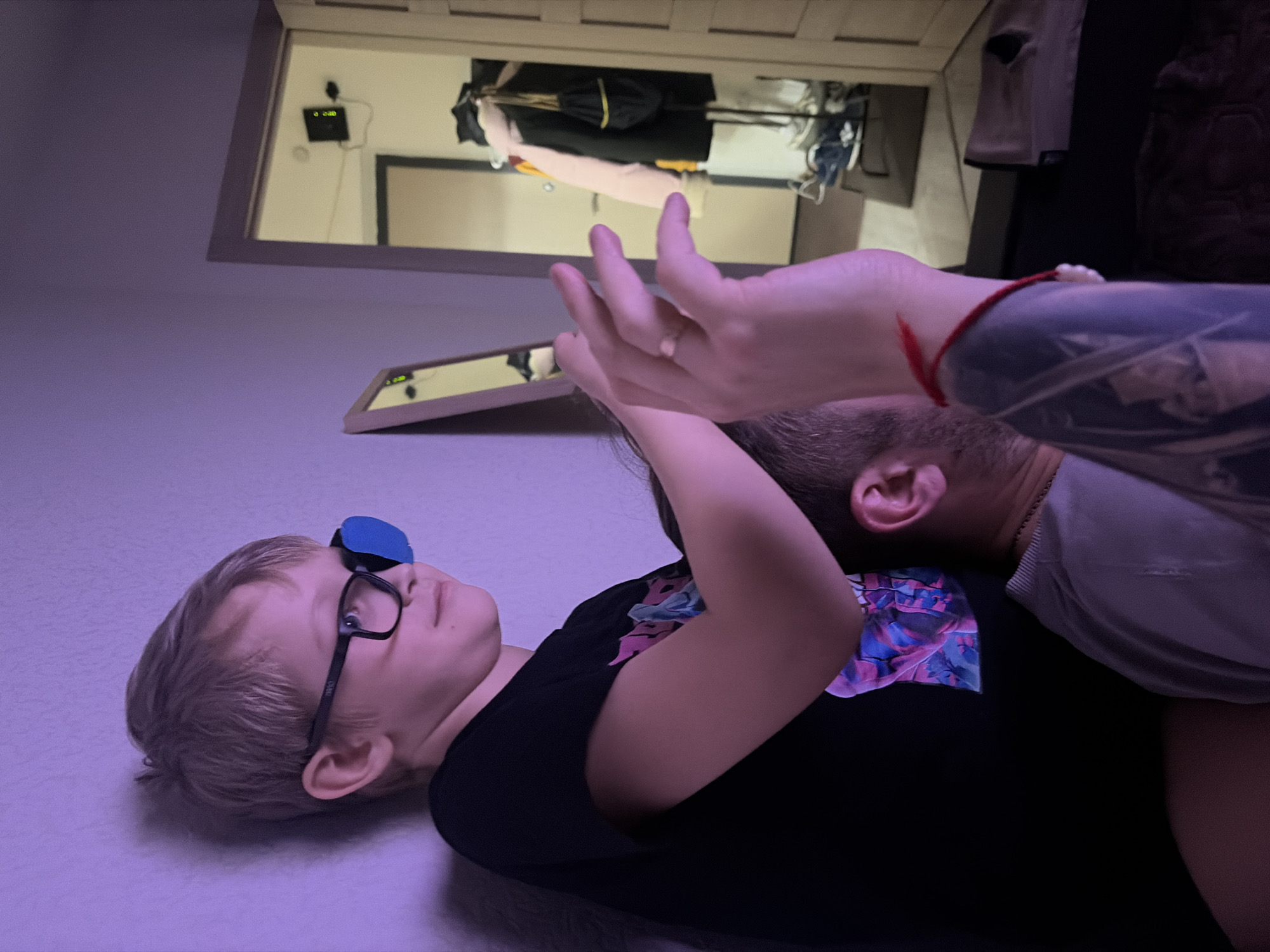 A distorted, low-angle indoor shot depicts a child wearing glasses and an eye patch lying on what appears to be a bed. An adult is beside the child, possibly embracing them. The adult has tattoos on their arm and is wearing a red bracelet. The background includes a doorway and reflections in what may be mirrors or frames. The location is undetermined, and there is no evidence linking it to Saint Petersburg, Russia.