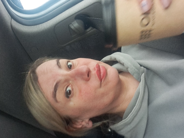 A fair-skinned woman with blonde hair and light eyebrows is angled from below, wearing a grey hooded sweatshirt. She’s holding a brown paper cup with a black lid; printed text is visible on the cup. The background appears to be the interior of a car with a window visible at the top left. The woman's expression suggests she is taking a selfie.