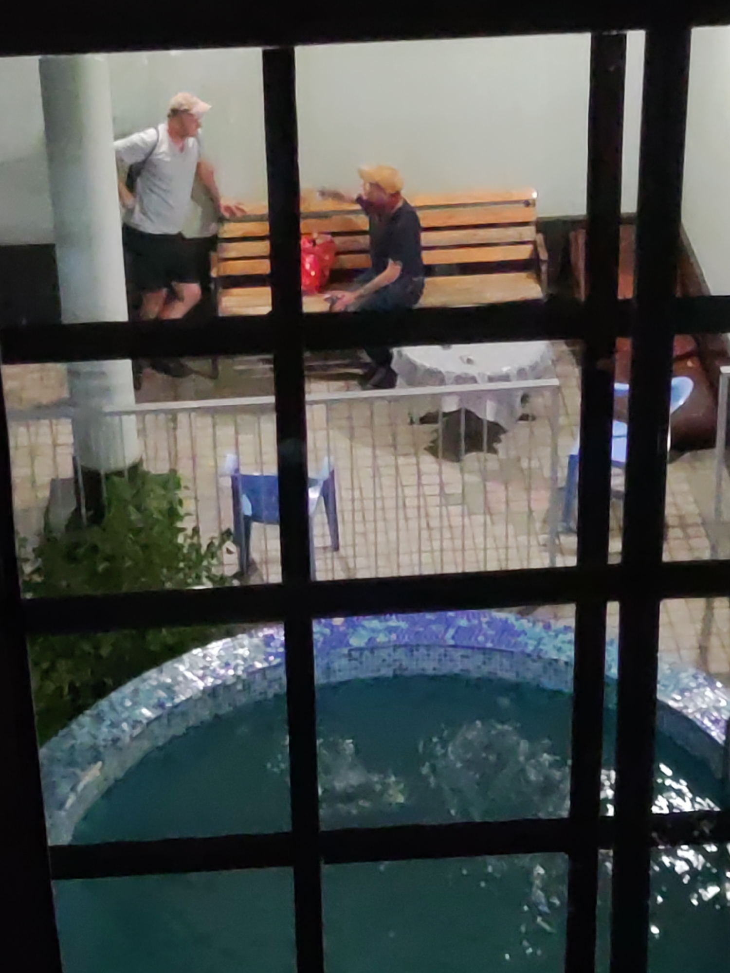 A dimly lit, outdoor patio area is viewed through a window with dark bars. In the background, two men wearing baseball caps are conversing while seated on a wooden bench. One man on the left wears a light-colored polo shirt and dark shorts. The man on the right wears a dark t-shirt and dark pants. A red bag sits on the bench between them. In the mid-ground, a round table covered with a white cloth is visible, along with several blue chairs. Lush green foliage is also present, near a light-colored pillar. In the foreground, a portion of a circular swimming pool with blue tiled edging is filled with bubbling turquoise water. The scene appears to be a hotel or residential complex in Tashkent, Uzbekistan.