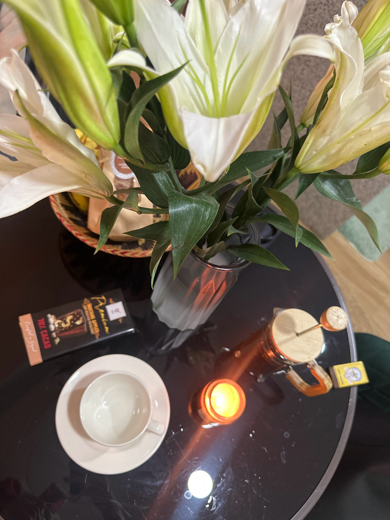 The image shows a close-up, high-angle view of a black, reflective table in Lalitpur, Nepal. On the table, there is a bouquet of white lilies in a glass vase. To the left of the lilies, there is a woven basket containing what appears to be packaged goods. In front of the basket, a dark chocolate bar with the text "70% CACAO" is visible. Further to the left and slightly below, a white teacup and saucer are placed. In the center-right of the image, a glass French press is positioned next to a small, lit candle in a glass holder. A small yellow box, possibly of matches, is placed near the French press. The lighting suggests an indoor setting with artificial light sources, as evidenced by the reflections on the table and the glow of the candle. There are no people visible in the scene.