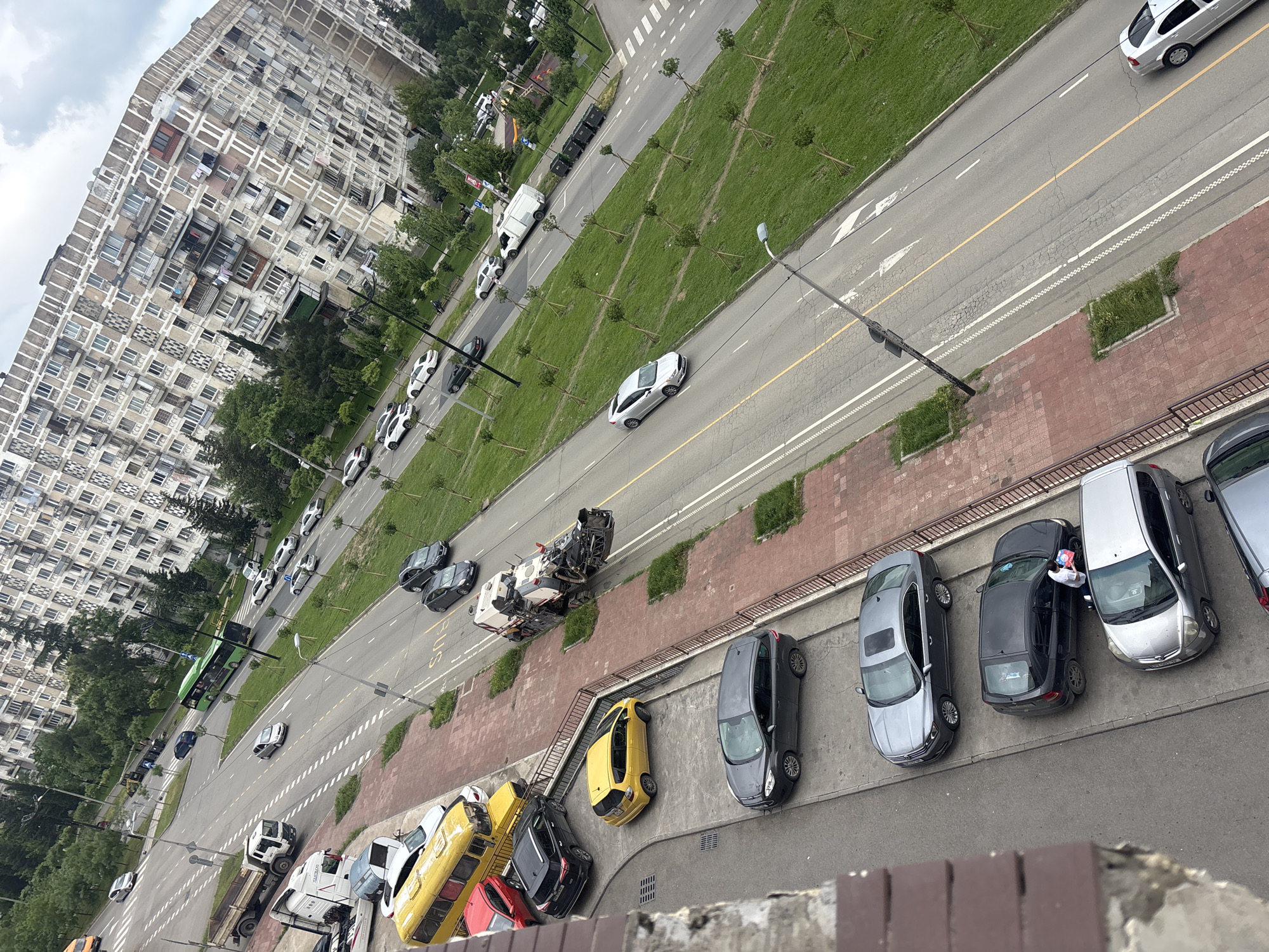 The image is taken from an elevated perspective, likely from a building overlooking a city street in Tbilisi, Georgia. The architecture includes a large apartment complex, with concrete balconies, typical of Soviet-era construction. The street below has multiple lanes and is separated by a grassy median strip with young trees.
Vehicles are present throughout the image, including cars parked along the sidewalk, several vehicles moving along the road, including a white van and a silver car. Also, near the sidewalk, there is what appears to be a pile of construction or demolition debris, likely from damaged vehicles. A person appears to be interacting with one of the cars parked along the sidewalk.
The environment includes a cloudy sky overhead and greenery in the median strips and around the buildings. The sidewalk is marked with red brick along the street. The overall scene depicts an urban environment with typical city features.