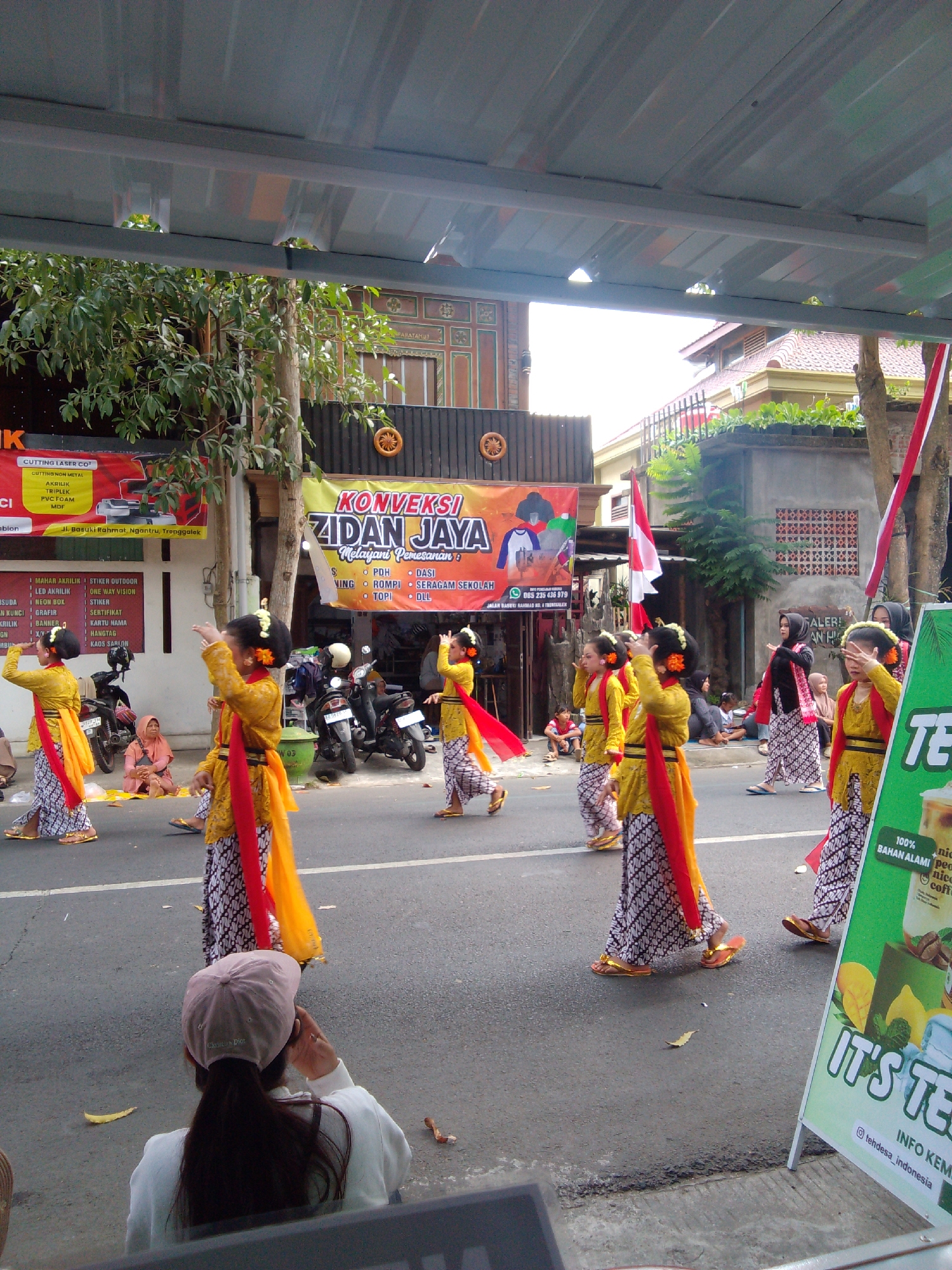 The image depicts a street scene in Trenggalek, Indonesia, likely during a cultural event or parade. Several young girls dressed in traditional yellow and orange attire, complete with elaborate hairstyles and red sashes, are performing a dance on the asphalt road. Their patterned skirts and golden slippers suggest a formal presentation. In the background, a banner with Indonesian text, including "ZIDAN JAYA," is visible, indicating a business or organization. Various other people are present, including spectators seated on the roadside and others observing from the sidewalks. Motorcycles are parked along the street, and buildings with traditional Indonesian architectural elements can be seen. The overall atmosphere is festive and communal, capturing a moment of cultural expression within the urban setting.