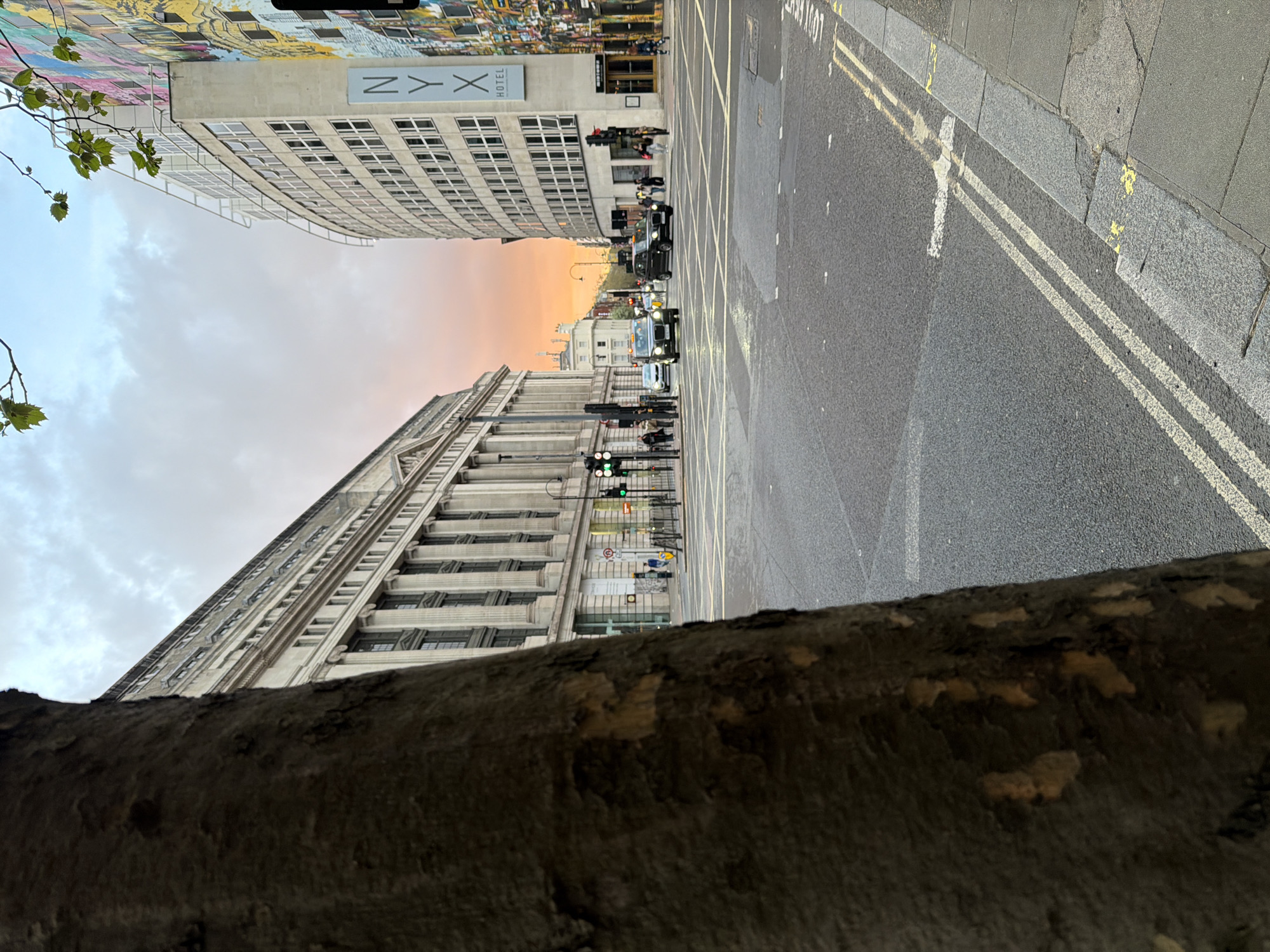 The image depicts a street scene in London, United Kingdom, with a distinct perspective. A tree branch occupies the foreground, partially obscuring the view. The street itself is visible, featuring a double yellow line marking. Buildings line both sides of the street, with one identifiable by the "NYX" sign on its facade. Several black taxi cabs are seen in the street. Pedestrians are present on the sidewalks. The sky above exhibits a warm orange hue, suggesting sunrise or sunset.