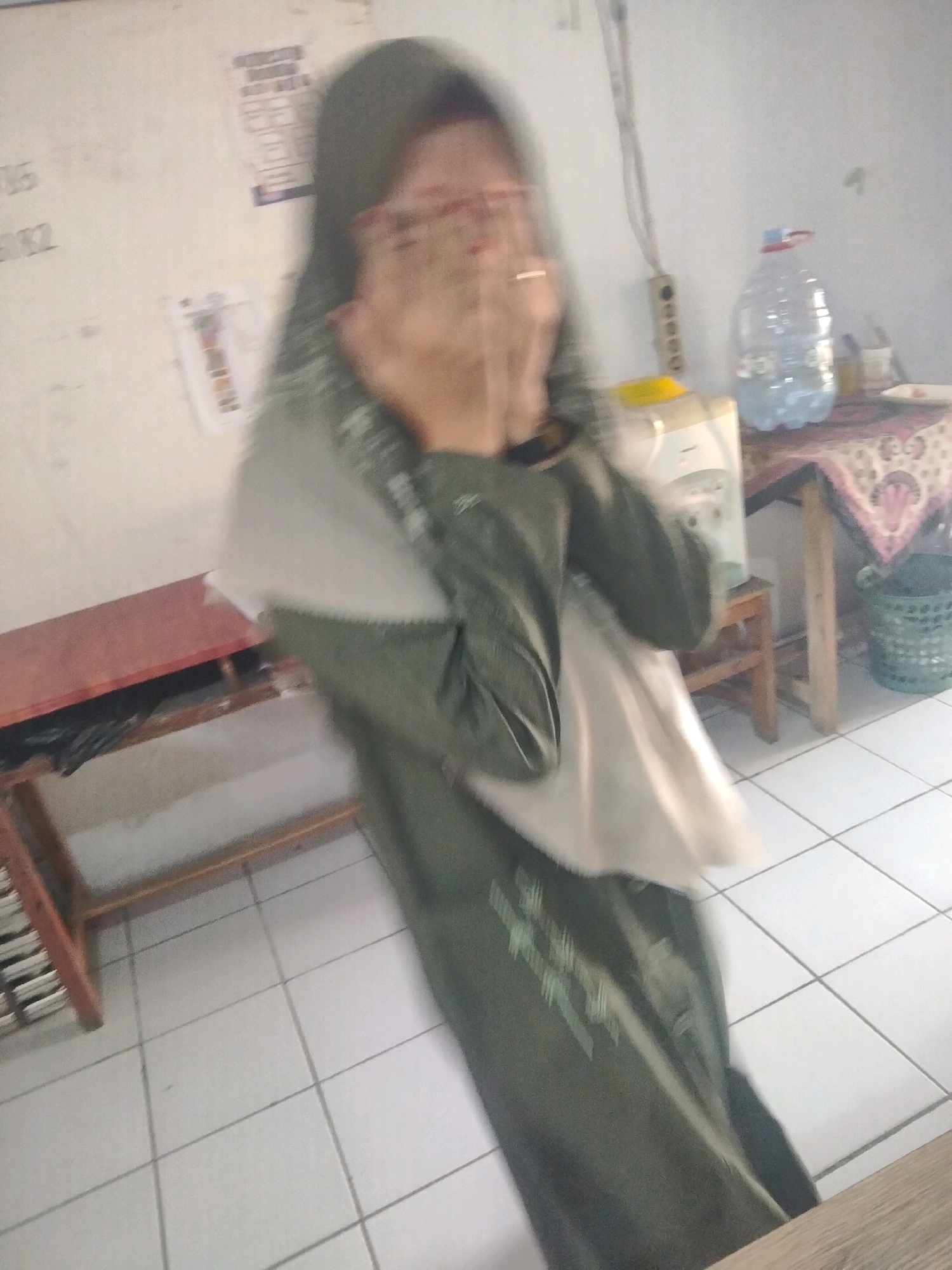 The image shows a blurry, full-body shot of a person wearing a dark green hijab and a matching long-sleeved dress with white accents. Their hands are covering their face, and they are wearing glasses. The person is standing in a room with white tiled floors and light-colored walls. To the left of the person is a wooden table with a red top and black trash bags underneath. In the background, there is a water dispenser, a plastic water bottle, a small table with a patterned cloth, and a green woven basket. There are papers or posters on the wall in the background. The lighting suggests it is daytime, possibly indoors. The image is distorted, making it difficult to discern specific details about the person or the surroundings. No specific Suka Sari, Indonesia landmarks are visible.
