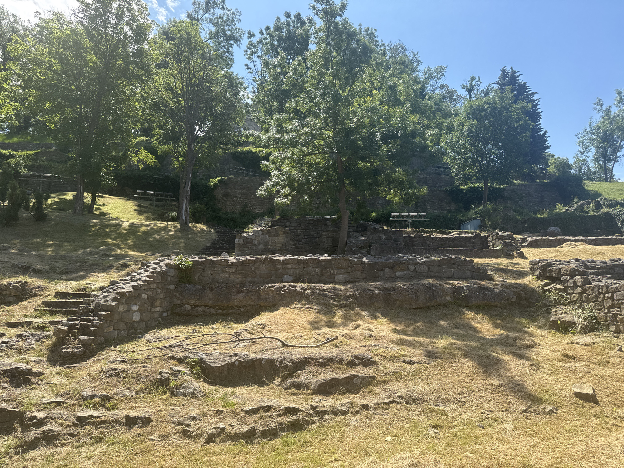 The image depicts an outdoor archaeological site on a sunny day. The foreground features ancient stone ruins, including a crumbling wall and a set of stone steps leading upwards. The ground is covered in dry, yellowish grass, with scattered rocks and a fallen branch.
In the background, lush green trees stand tall on a hillside dotted with more stone structures and what appear to be remnants of walls and terraces. Some wooden benches are visible among the trees, suggesting this is a historical site that is open to visitors. The sky is a clear, bright blue, indicative of fair weather. The dappled sunlight and shadows suggest it is late morning or early afternoon. The overall impression is of a peaceful, sun-drenched historical location, possibly a Roman or medieval ruin, nestled in a natural setting. No people are visible in the image.