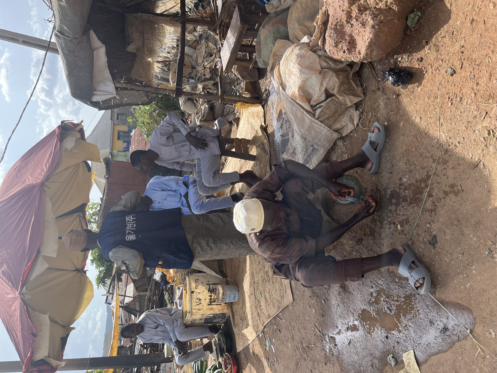 In Jos, Nigeria, a busy outdoor market scene unfolds under a worn-out red and tan umbrella. Several men are present, some seated, others crouched. One man, wearing a plaid shirt and pants with a white cap, is actively engaged in processing something on the ground. Another man in a blue long-sleeved shirt and pants sits on a wooden stool, while a third man, also in a light blue outfit, is seated nearby. A fourth man, dressed in a grey traditional outfit, sits on a white crate. In the background, market stalls with corrugated iron roofs and makeshift shelters are visible, suggesting a vibrant commercial hub. The ground is a mix of dirt and puddles, with various sacks and rocks scattered around. The image captures a moment in the daily life of this Nigerian marketplace, with a focus on human activity and the surrounding environment.