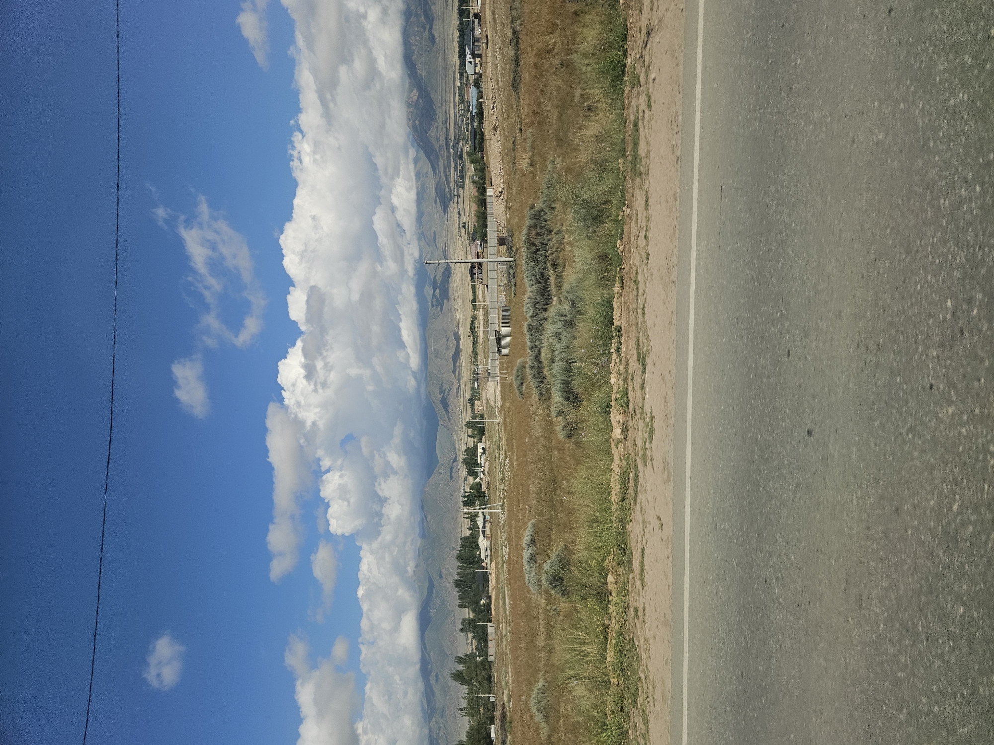 The image depicts a wide-angle view of a rural landscape, likely in or near Ornek, Kyrgyzstan, under a bright blue sky with scattered white clouds. In the foreground, a portion of an asphalt road with a white line is visible, suggesting it is a roadway. Beyond the road, a dry, grassy field stretches out, dotted with sparse, low-lying bushes and taller, dry grass. In the mid-ground, a small village or settlement is nestled amongst trees, with several buildings and structures visible. Power lines and utility poles are also present, indicating infrastructure. In the background, a range of mountains forms a natural backdrop, with some of the peaks lightly shrouded in clouds. The overall impression is one of a vast, open, and somewhat arid rural environment. There are no people visible in the image.