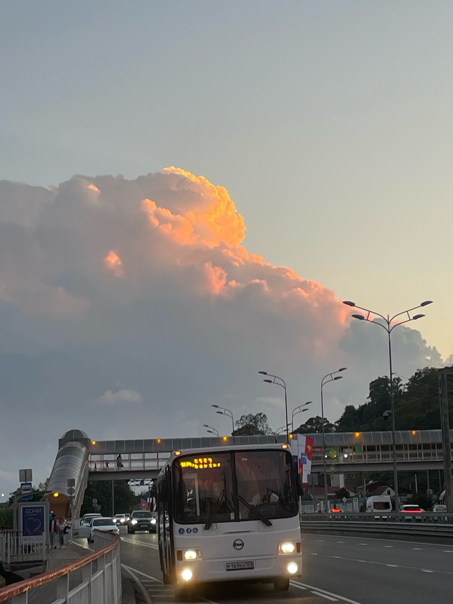 The image depicts a street scene in Sochi, Russia, at dusk. A large bus, displaying yellow illuminated text on its front, is driving on a multi-lane road. In the background, an elevated pedestrian bridge with glass enclosures and yellow lights spans across the road. To the left of the bridge, a ramp leads up to it, and a sign with the word "СОЧИ" is visible. Several cars are on the road, and people are walking on a sidewalk near the pedestrian bridge. The sky above is filled with dramatic clouds, some of which are illuminated with warm, orange and pink hues, suggesting the setting sun. Trees and buildings are visible on a distant hillside.