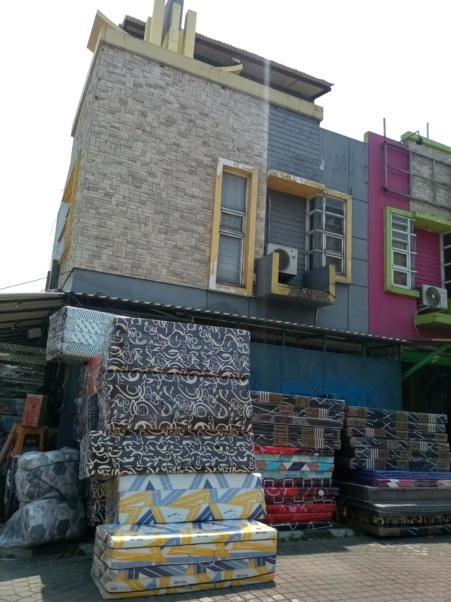 In Bekasi, Indonesia, a street-level outdoor view captures a building with multiple levels and colorful facades, predominantly featuring a beige stone-patterned lower section and a grey upper section. To the right, a vibrant pink facade with green trim and windows is visible. In front of the building, a large quantity of mattresses are stacked on the pavement, some wrapped in clear plastic and others with patterned coverings. The mattresses are arranged in stacks of varying heights and sizes. A blue metal gate is visible behind some of the mattresses. The scene appears to be a street-level storefront or warehouse displaying mattresses for sale. The sky is overcast and bright.