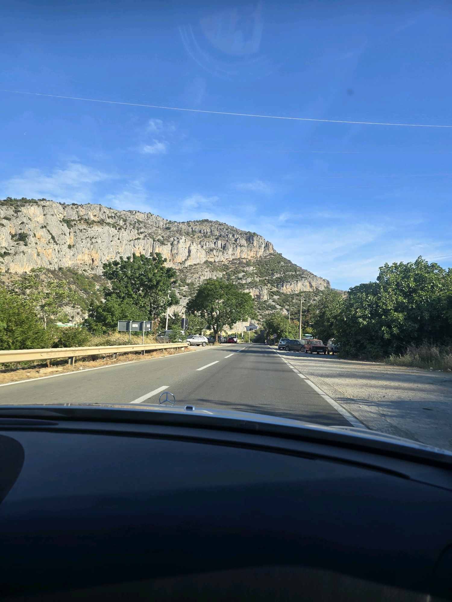 The image depicts a view from inside a vehicle, looking forward on a paved road. The road is bordered by a guardrail on the left and a grassy shoulder on the right. Beyond the guardrail, there is a steep, rocky mountain range that dominates the background. Lush green trees and vegetation grow on the lower slopes of the mountain and along the roadside. Several cars are parked or driving on the road, and some appear to be parked on the shoulder. The sky is bright blue with a thin white contrail from an aircraft visible. The scene suggests a journey through a scenic, mountainous region.