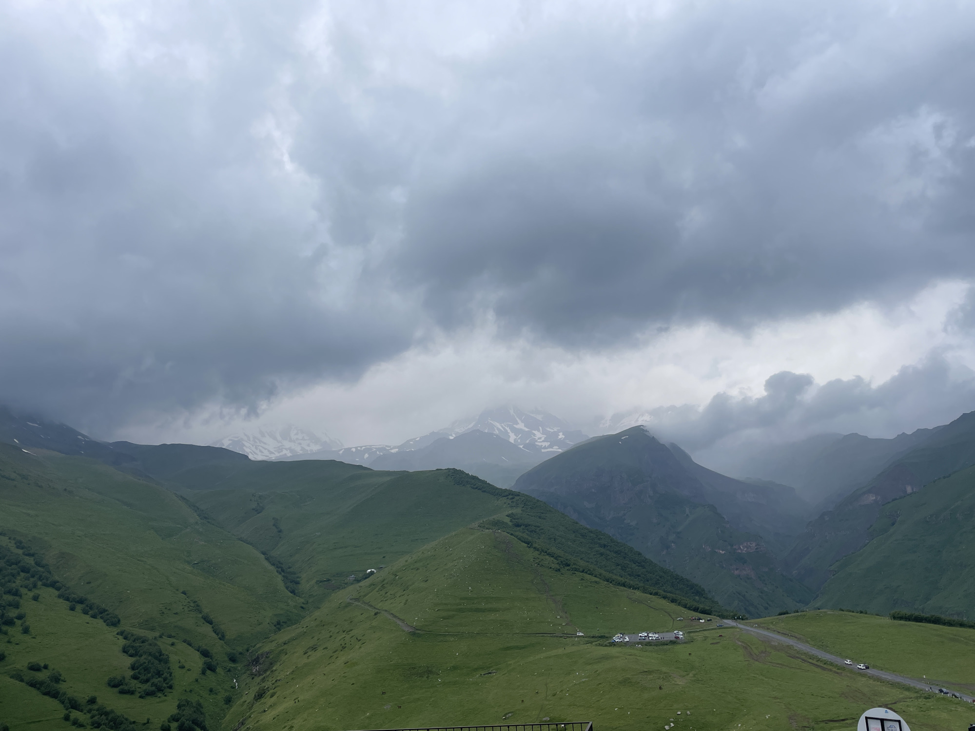 The image depicts a vast, mountainous landscape under a heavily overcast sky. Rolling green hills dominate the foreground and midground, with a winding dirt road visible on one of the slopes, accompanied by a parking area with several white vehicles. In the distance, larger, more rugged mountains rise, some capped with snow, shrouded in mist. The overall atmosphere is somber and natural, suggesting a remote, elevated location. This scene is characteristic of the high-altitude terrain surrounding Stepantsminda, Georgia, a town renowned for its dramatic mountain vistas, including the iconic Mount Kazbek.