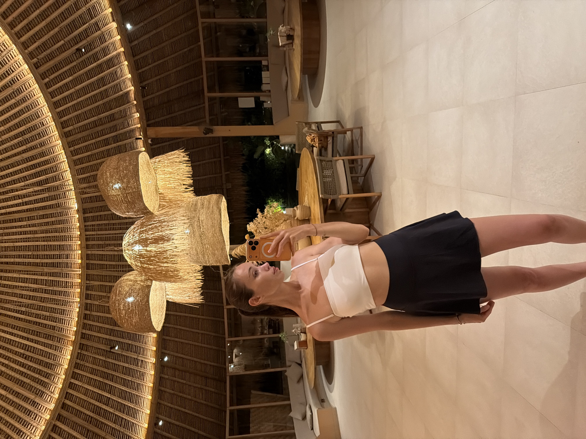An interior view depicts a female subject in a lounge or dining area located in the city of Unknown, Indonesia. The overhead ceiling is a domed structure constructed from natural wood and thatch materials, with visible concentric rafters and integrated perimeter lighting. Three large, conical pendant lights, fabricated from woven natural fibers with decorative fringes, hang suspended from the ceiling. The floor consists of large, light-beige square tiles.
The sole individual, a female with dark hair pulled back into a ponytail, stands near the center foreground. She is dressed in a white strappy crop top and a black mini-skirt. Her right hand holds an orange-cased smartphone at arm's length, angled towards her, indicating a self-portrait capture. A thin bracelet is visible on her left wrist, with her arm positioned at her side.
In the background, a lounge area features a curved, built-in sofa with light-colored cushioning, situated against a wall and fronted by a large, low, circular wooden coffee table. To the right, two round wooden tables are present, surrounded by chairs constructed from woven material with light-colored cushions. Several tables are adorned with vases containing dried botanical arrangements or small potted plants. The perimeter of the space is defined by large glass panels that look out onto a dark exterior environment, where artificial lighting illuminates distant foliage. The scene is lit by interior ambient lighting and the glow from the pendant fixtures.